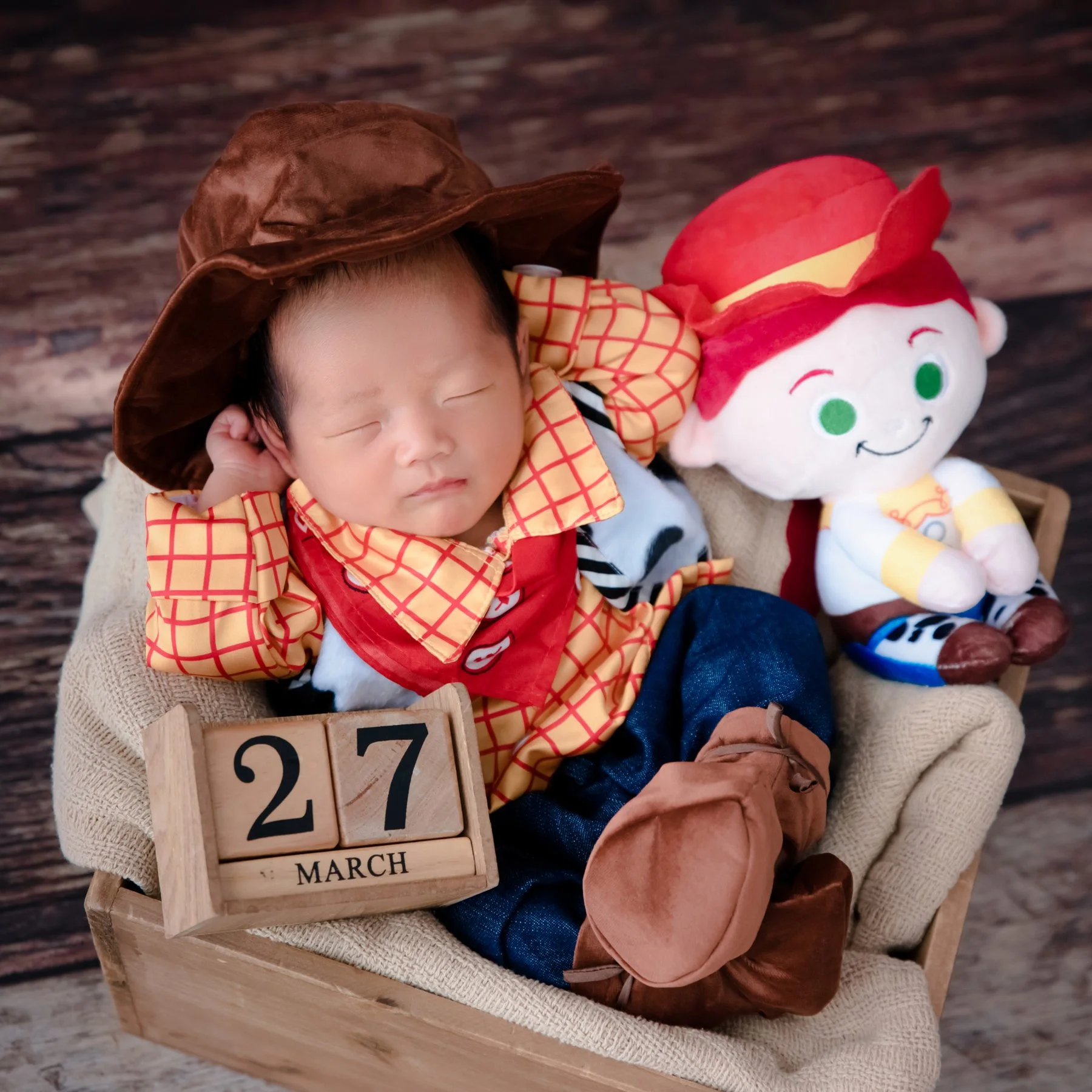 Sleeping newborn in a Woody cowboy outfit and brown hat seated beside a Jessie plush in a rustic basket