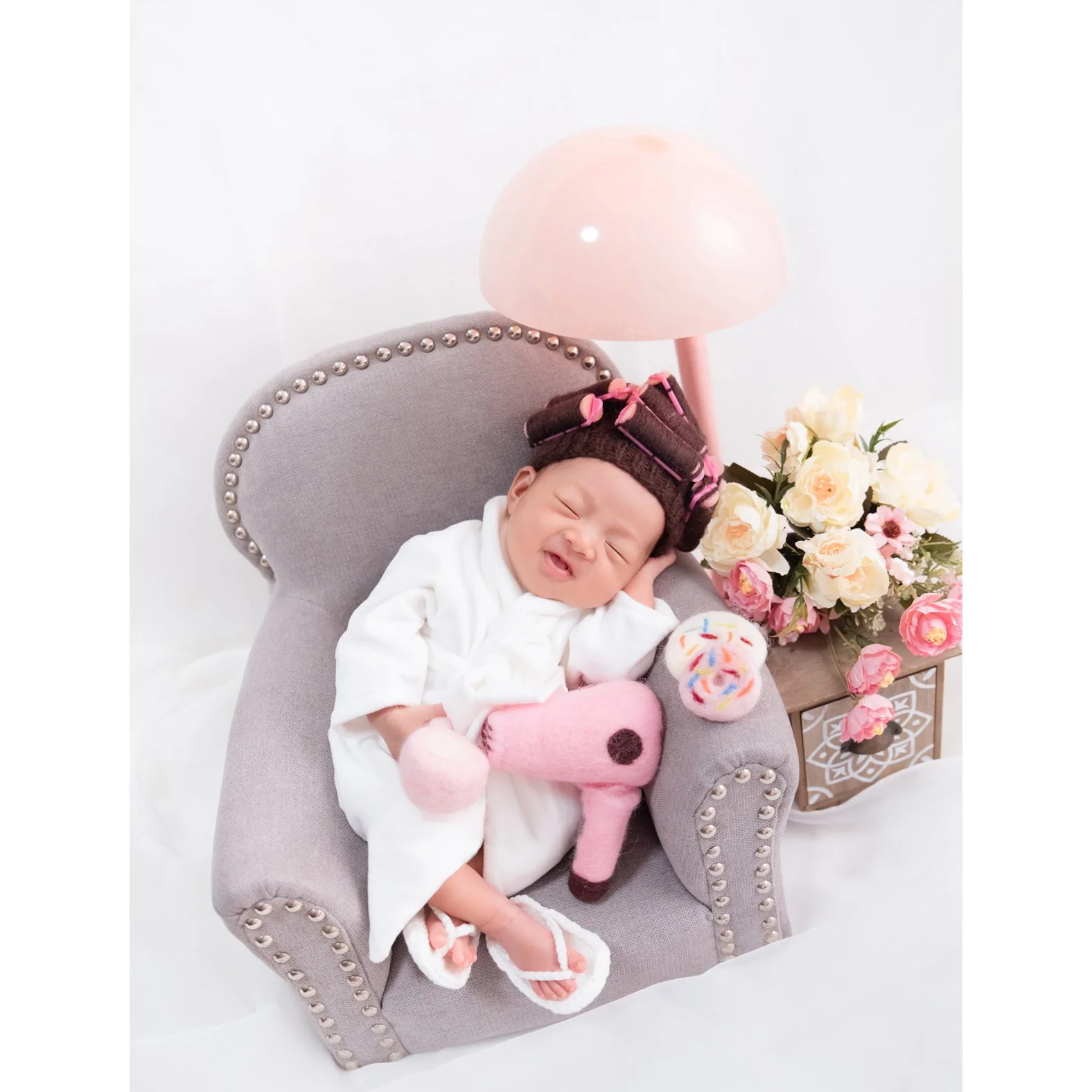 Smiling newborn in a white bathrobe and pink hair rollers reclining in a miniature grey armchair with a pink giraffe toy and flowers