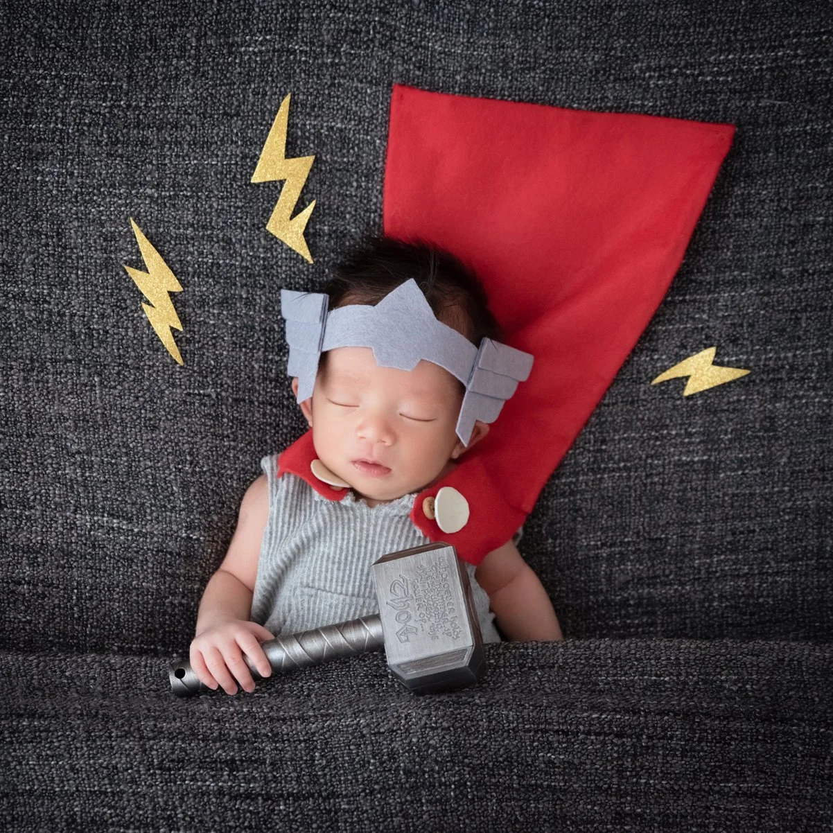 Sleeping newborn wearing a Thor helmet with a toy hammer across the front and a red cape behind