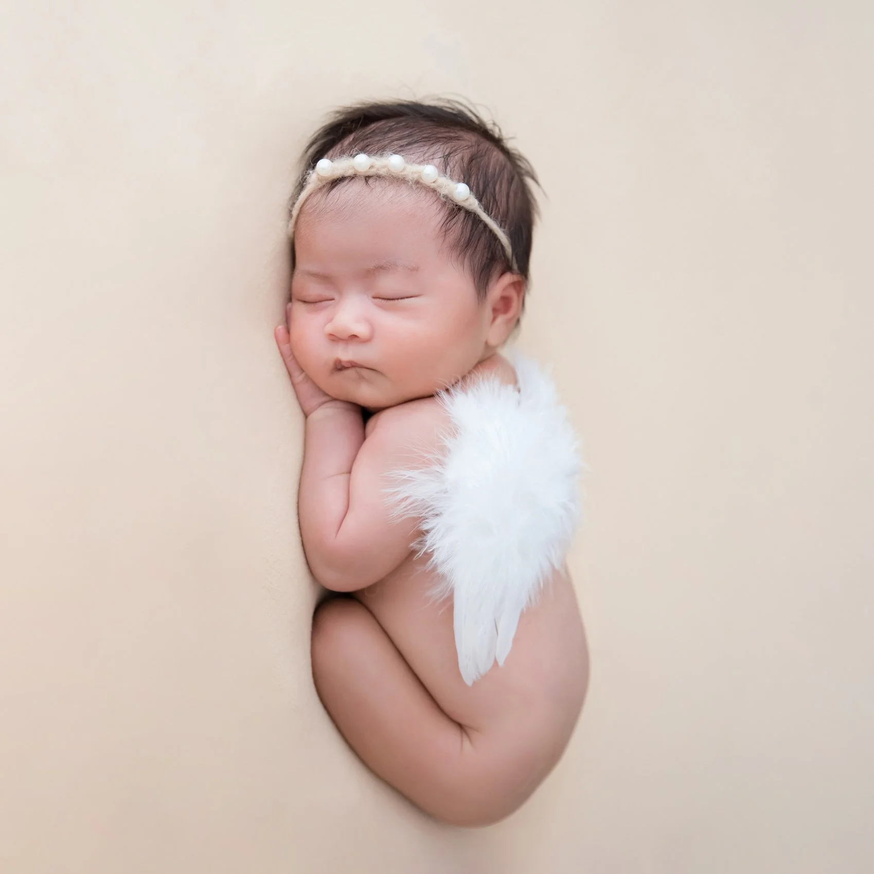 Sleeping newborn with a white headband and feathered angel wings curled on one side against a beige backdrop