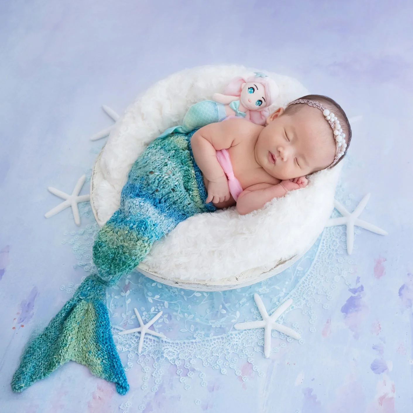 Sleeping newborn in a turquoise mermaid tail curled in a white basket with starfish props against lavender