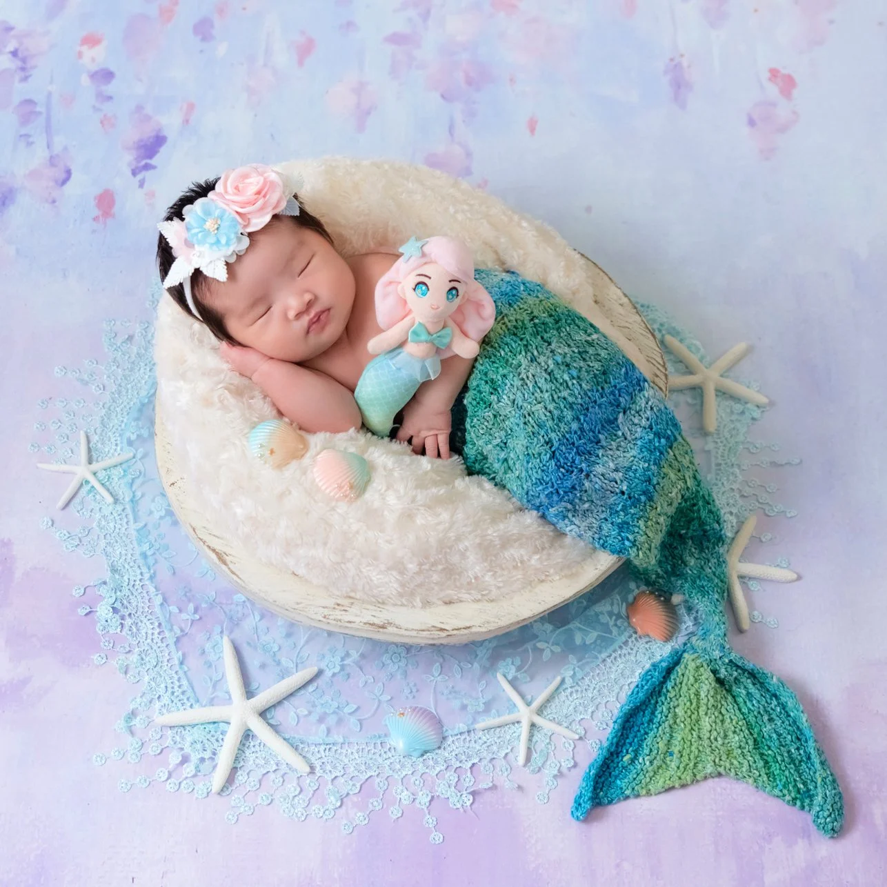 Sleeping newborn with a floral headband and turquoise mermaid tail nestled in a cream basket beside starfish