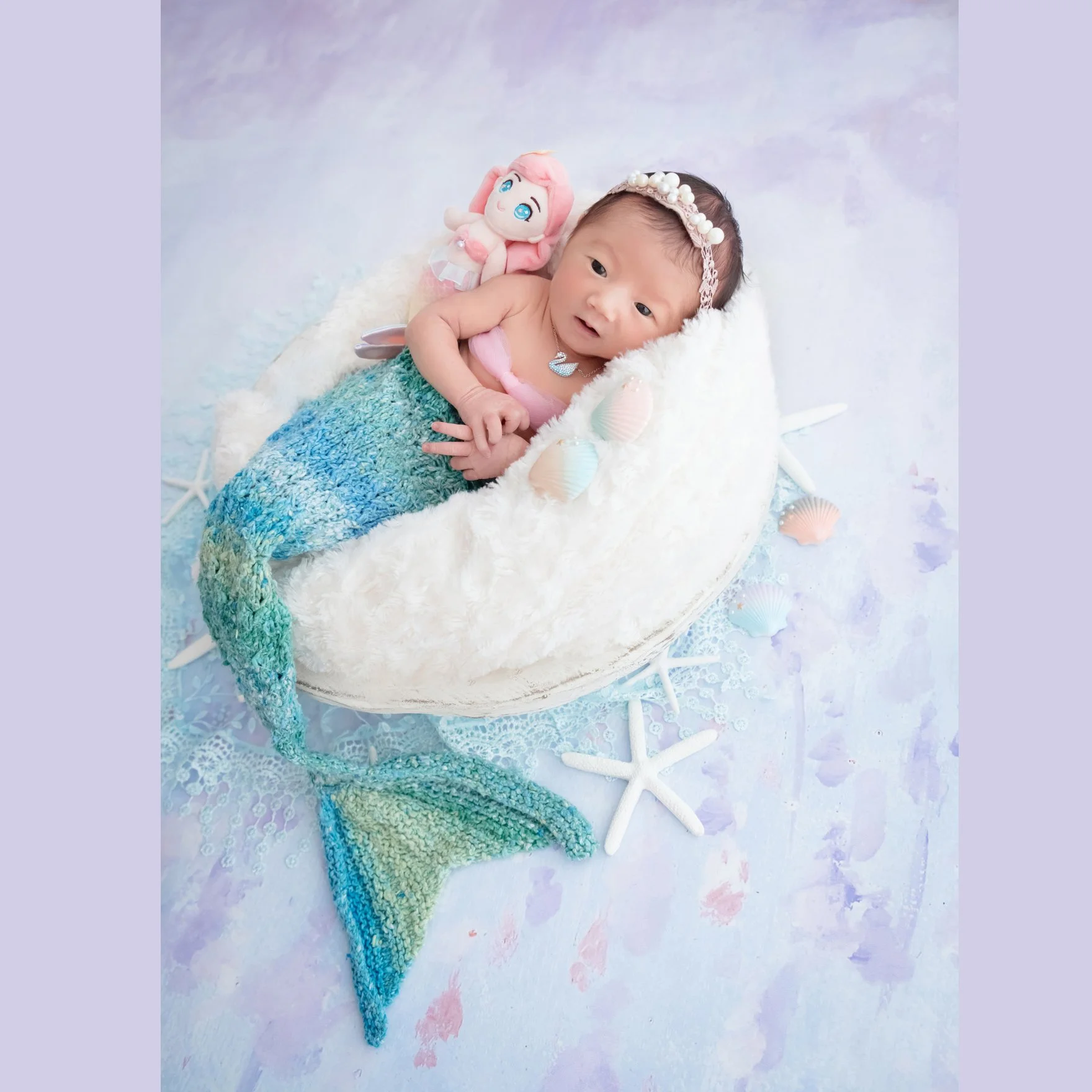 Awake newborn wearing a white headband and turquoise mermaid tail in a white basket beside a mermaid toy