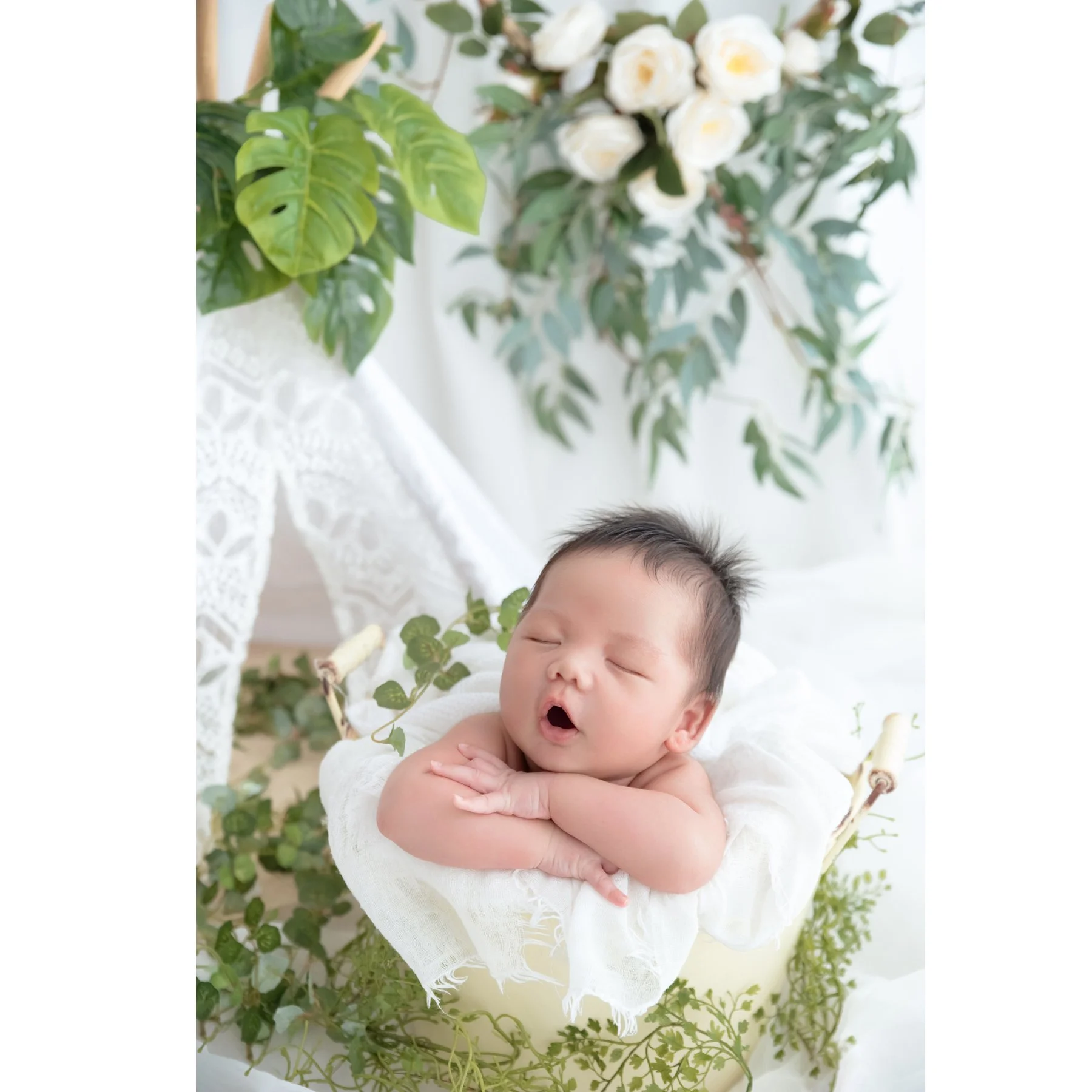 Yawning newborn resting the chin on folded arms in a white-lined basket beneath white flowers and green leaves