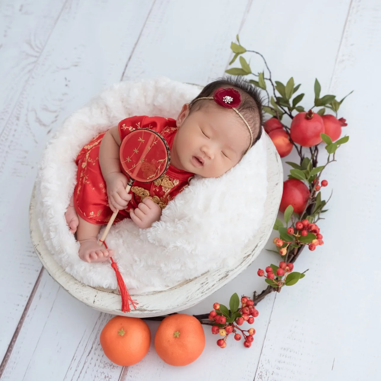 Sleeping newborn in a red outfit and matching headband curled in a white-lined basket beside oranges and red flowers on a pale wooden backdrop
