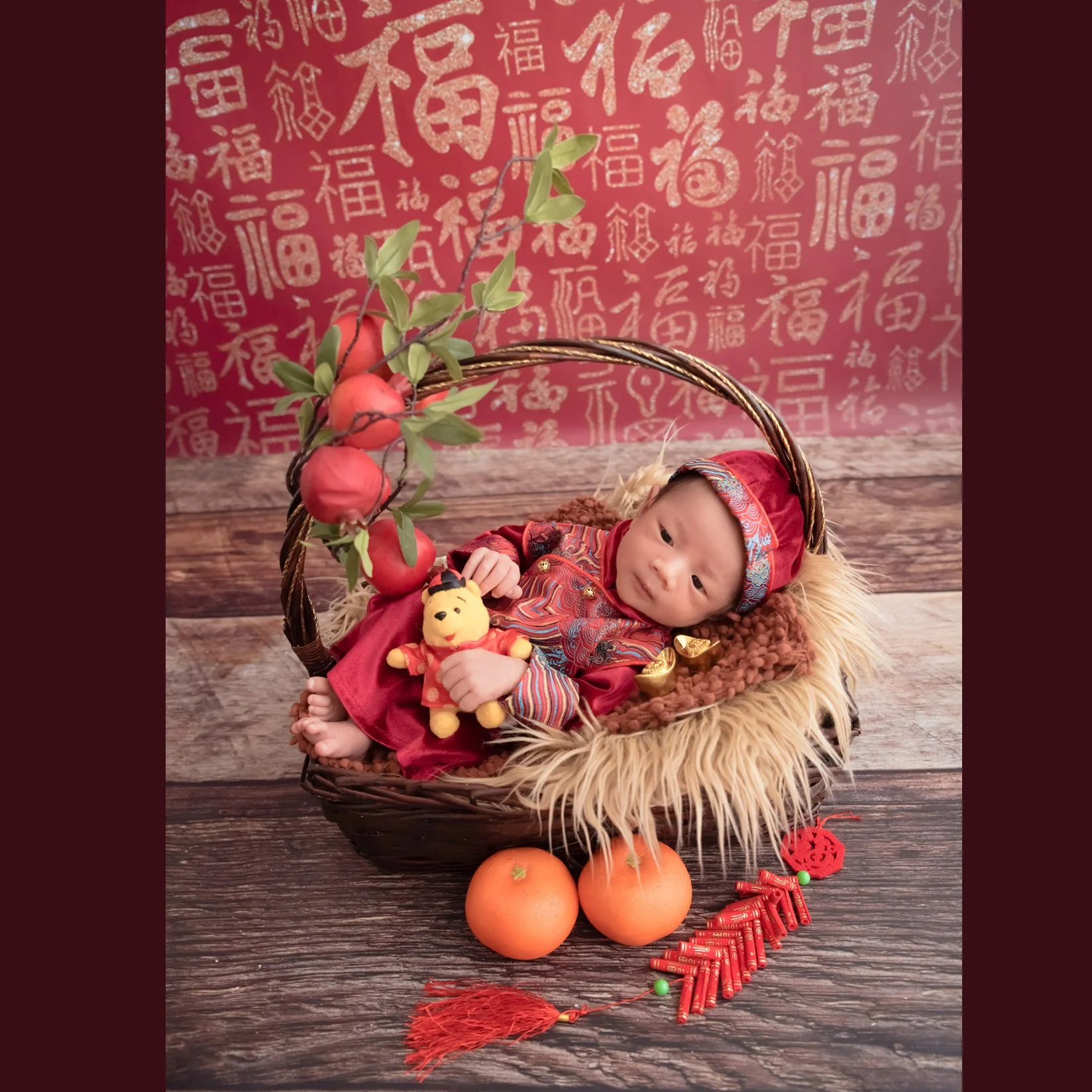Sleeping newborn in a red patterned outfit and matching headband reclining in a woven basket with a festive teddy bear, oranges and red Chinese New Year decorations
