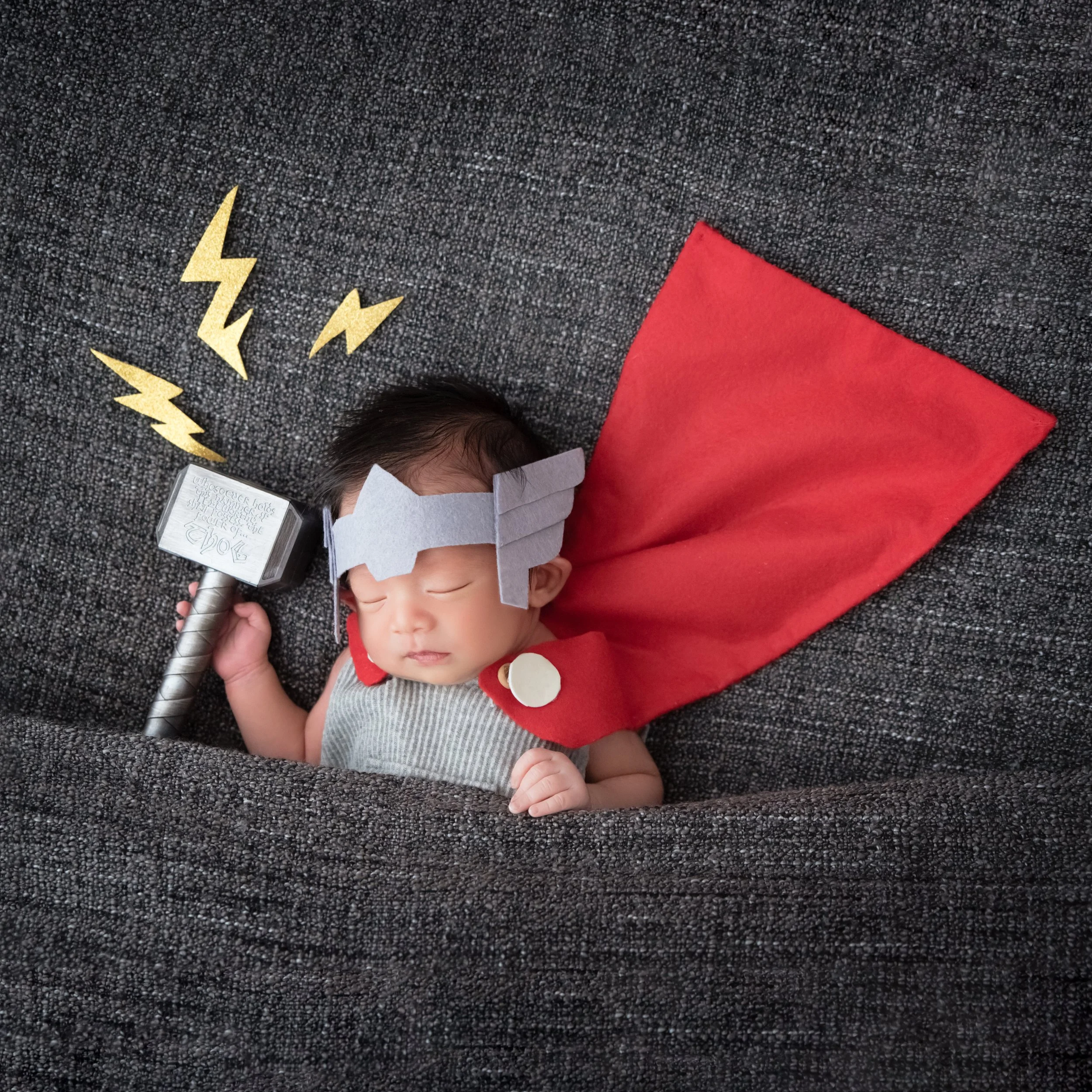 Sleeping newborn dressed as Thor with a raised toy hammer and red cape spread behind against a grey backdrop