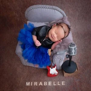 Sleeping newborn in headphones, a black jacket and blue tutu reclining in a miniature grey armchair beside a toy guitar and microphone