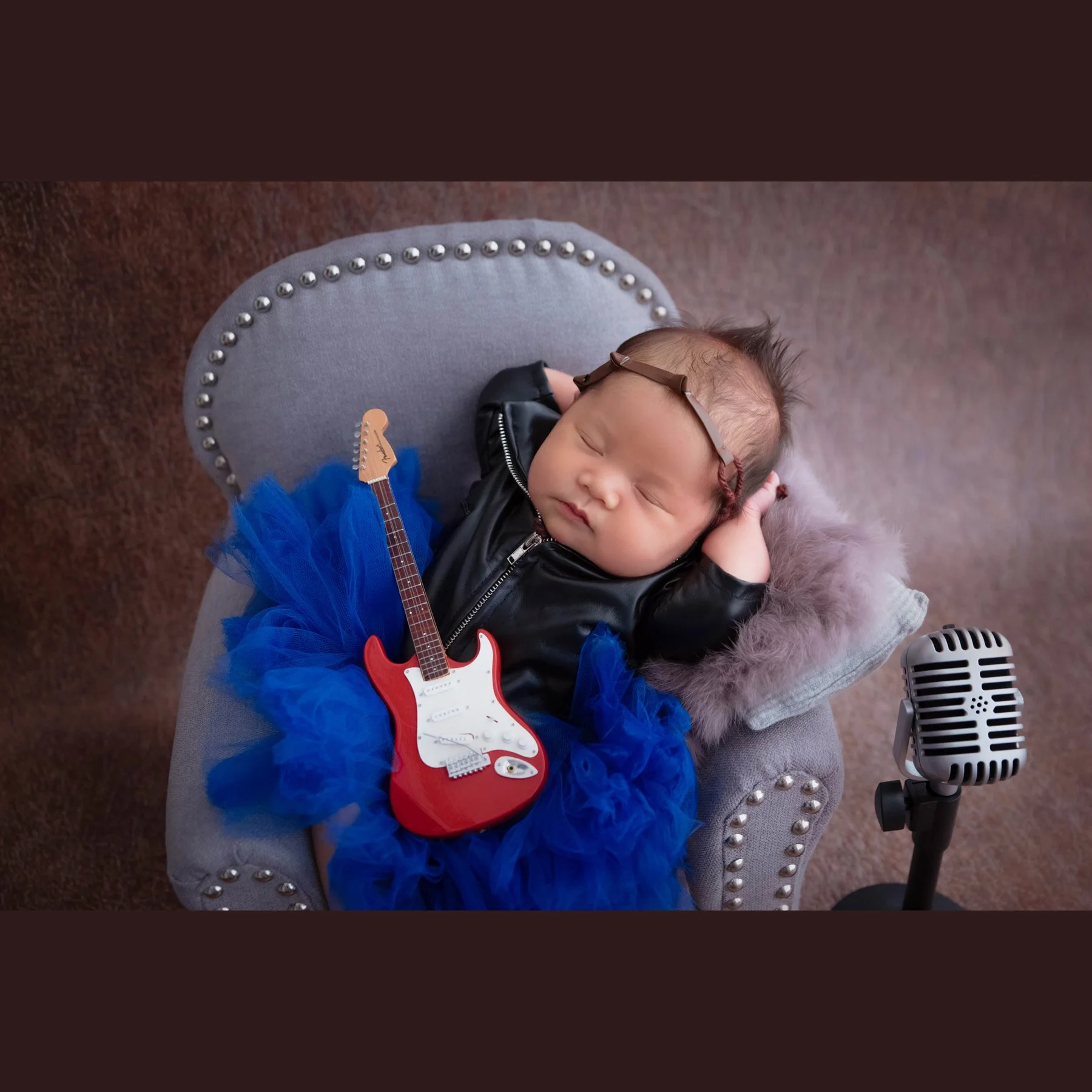 Sleeping newborn wearing headphones, a black jacket and blue tutu with a red toy guitar across the lap in a miniature grey armchair