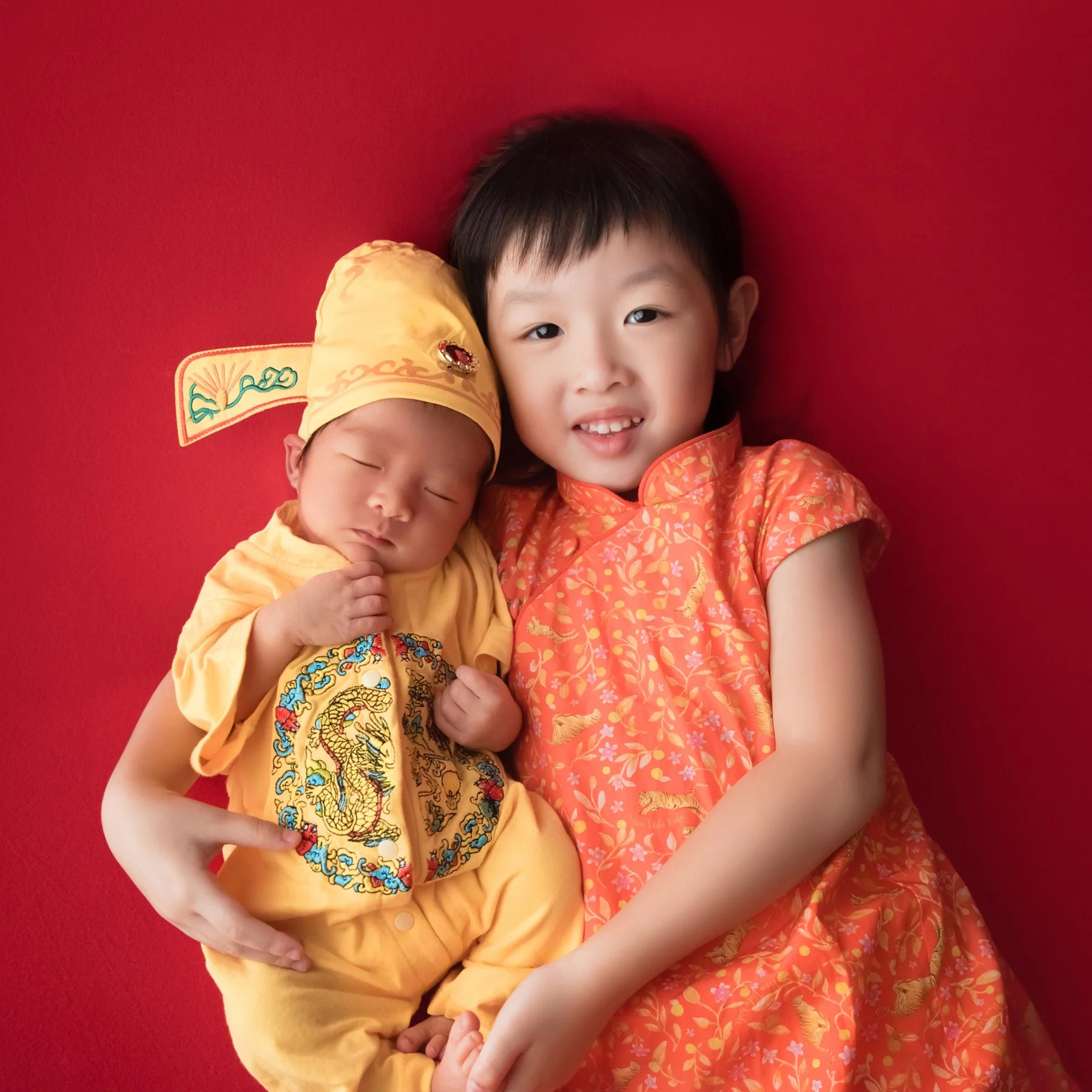 Smiling older sister in an orange cheongsam holding a sleeping newborn dressed in a yellow embroidered Chinese outfit and hat against a red backdrop
