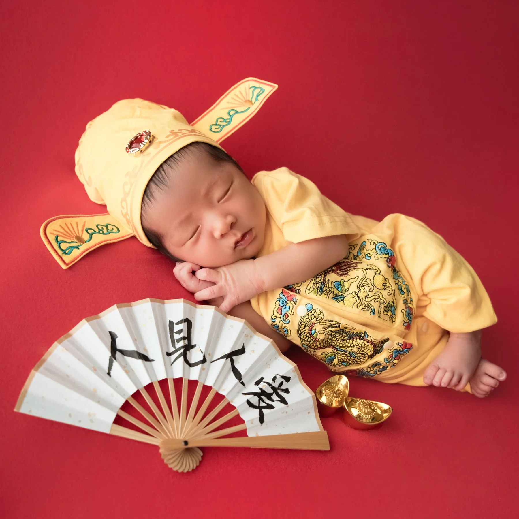 Sleeping newborn in a yellow embroidered Chinese outfit and hat lying on one side beside a calligraphy fan and toy gold ingots on a red backdrop