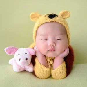 Sleeping newborn in a yellow Winnie the Pooh hat resting the cheeks on both hands beside a Piglet toy