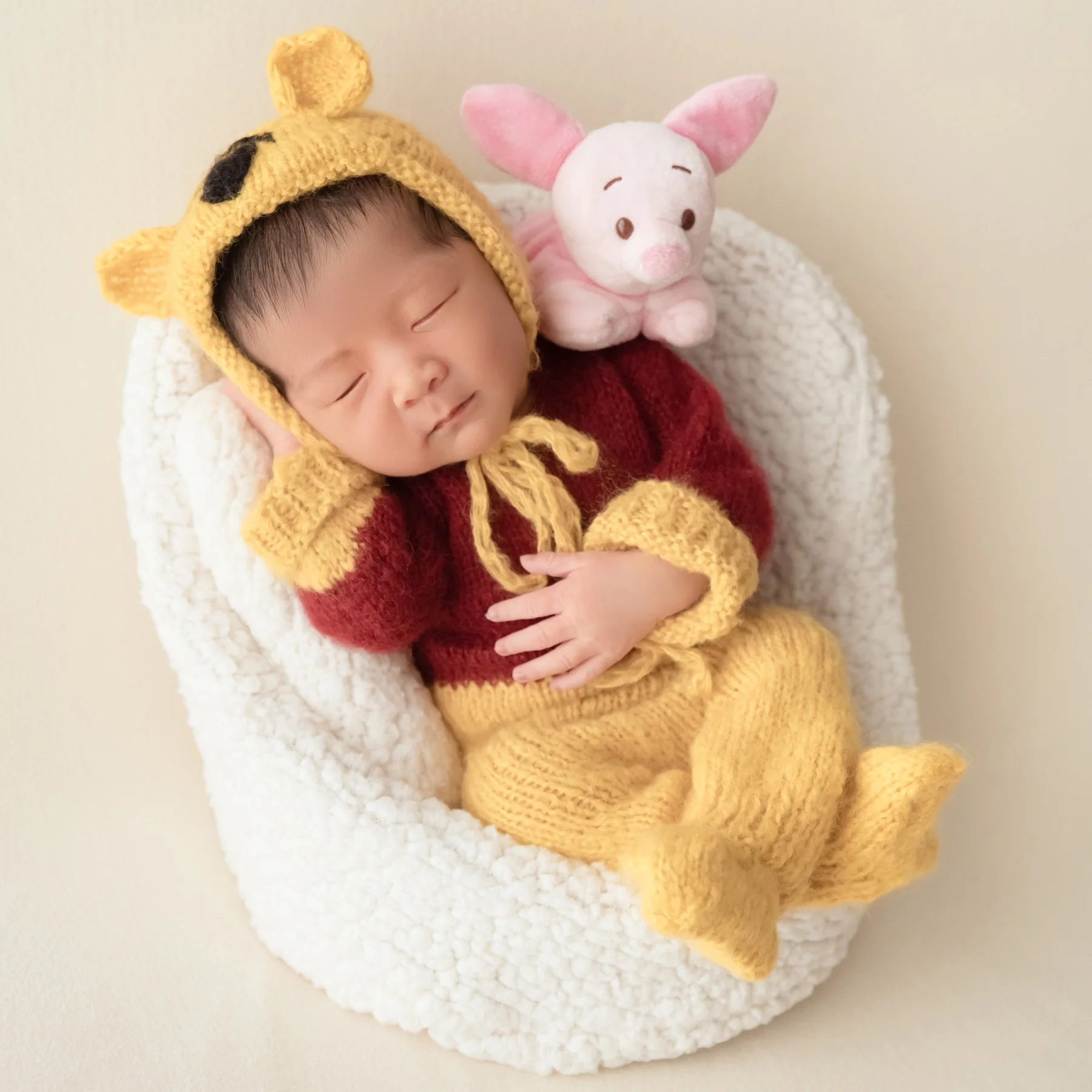 Sleeping newborn in a yellow Winnie the Pooh outfit with a Piglet toy tucked beside the head in a white basket