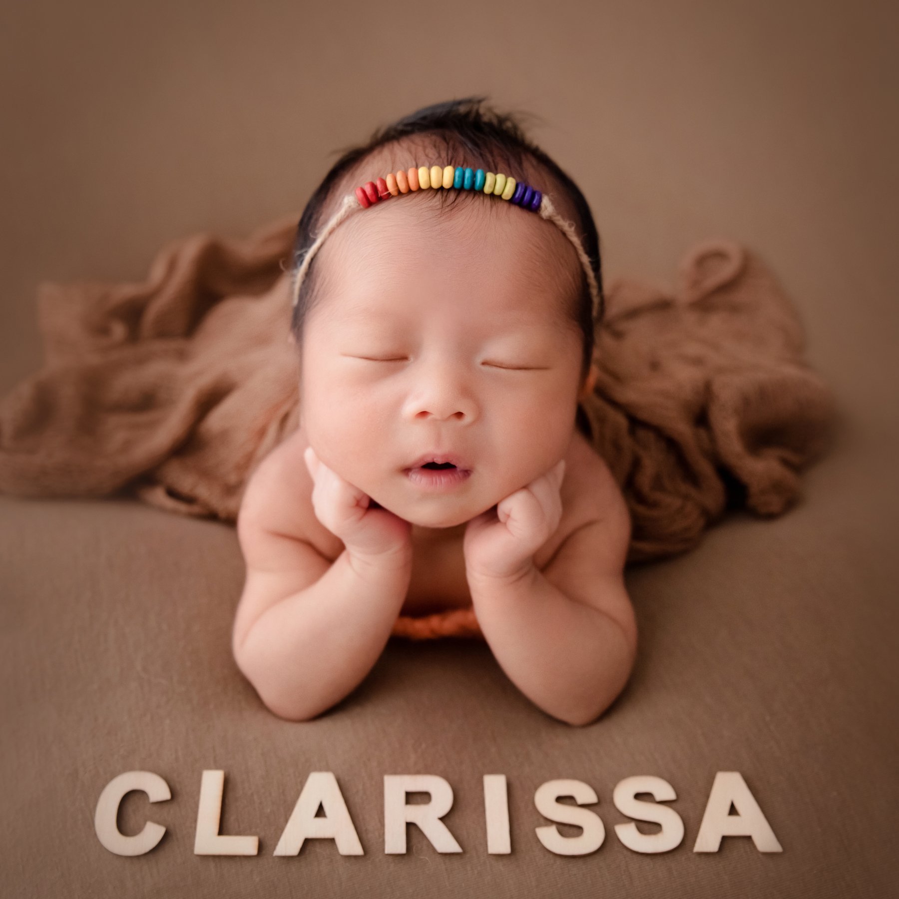 Sleeping newborn wearing a colourful headband resting both cheeks on hands on a plain brown backdrop