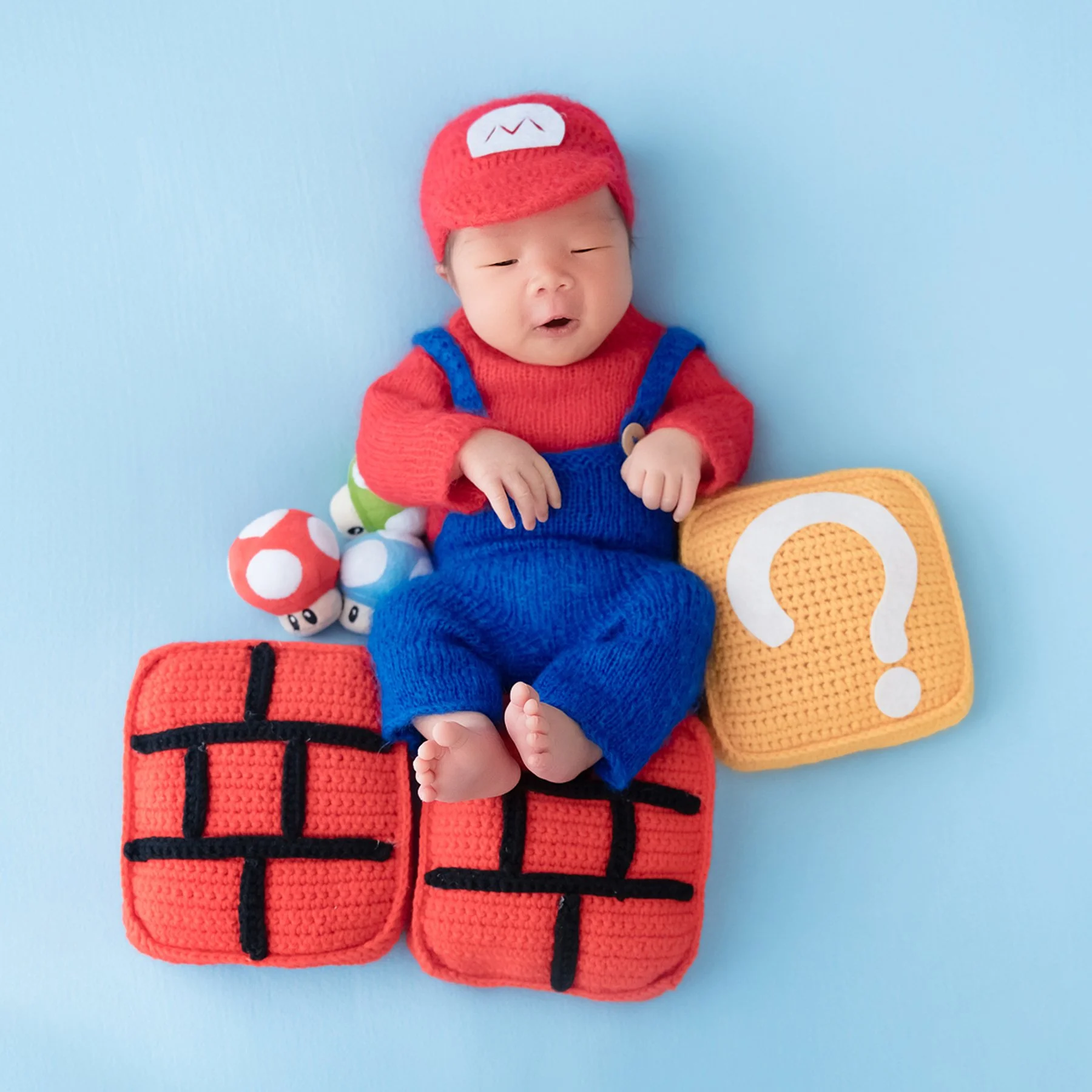 Newborn in a red Mario hat and blue knitted overalls resting on toy brick blocks beside a question block