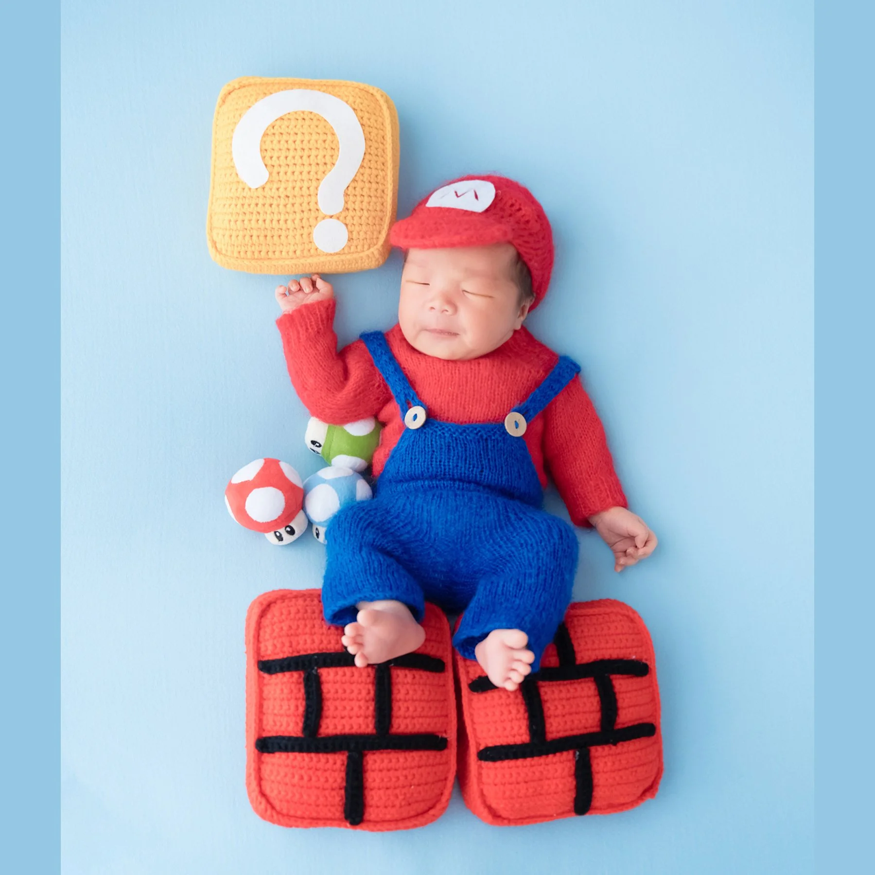 Sleeping newborn in a Mario outfit with one hand raised toward a knitted question block on a blue backdrop