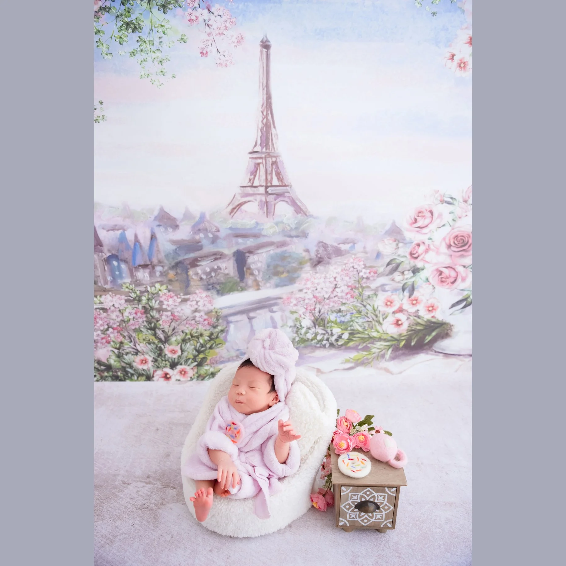 Awake newborn in a pink dress and matching headband reclining in a pink basket beside flowers and a vintage clock against an Eiffel Tower backdrop