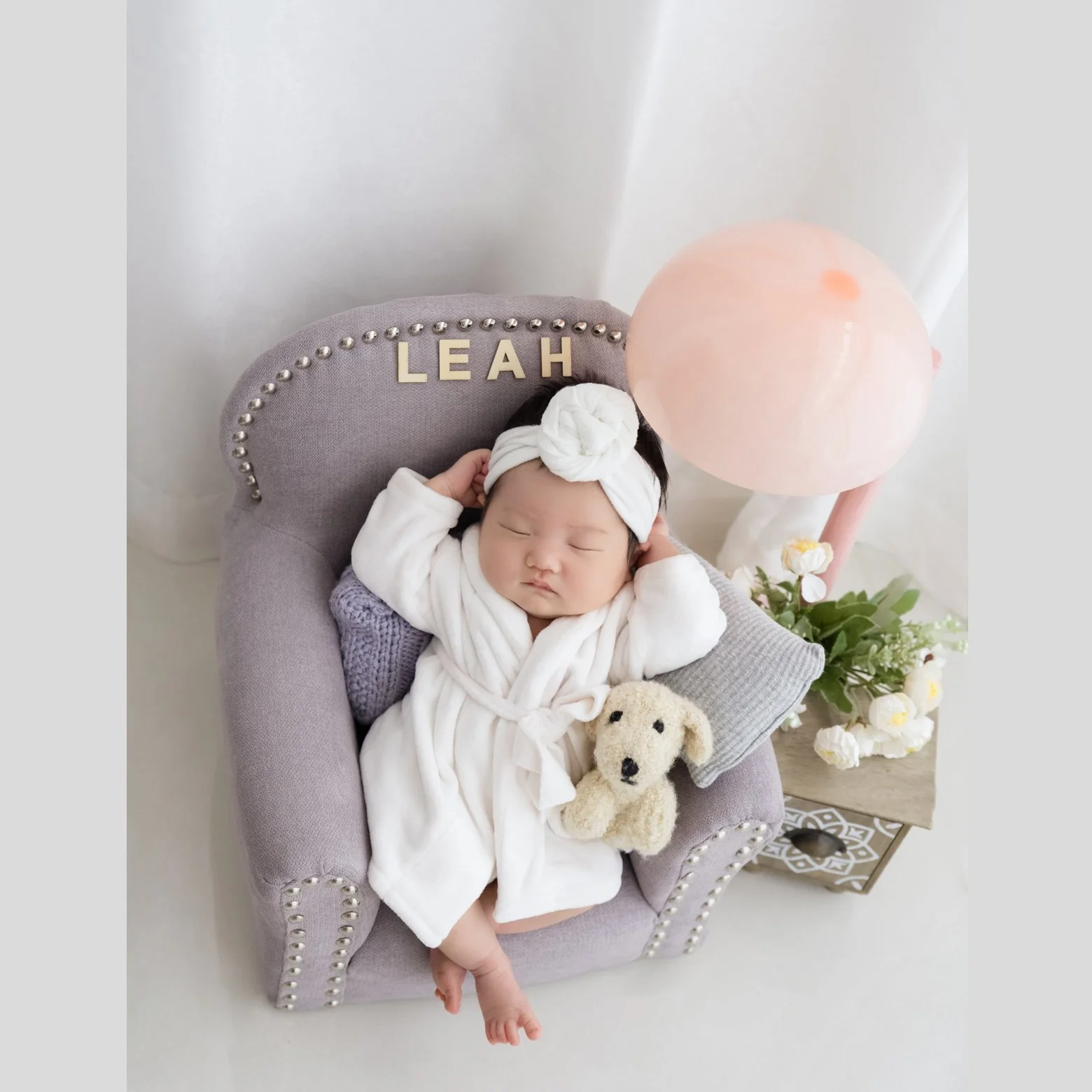 Newborn in a white bathrobe and knotted towel head wrap reclining in a miniature grey armchair with a cream teddy bear