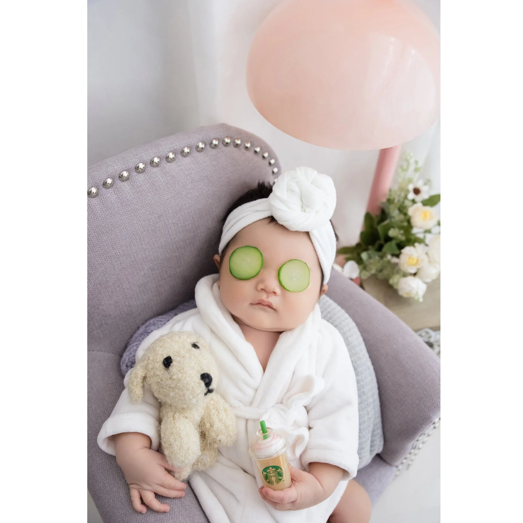 Close-up of a newborn in a white bathrobe and towel head wrap with cucumber-slice props over the eyes holding a cream teddy bear