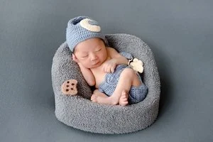 Smiling sleeping newborn in a grey knitted hat with a crescent moon and matching shorts curled in a soft grey basket beside a miniature cookie