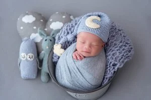 Sleeping newborn in a grey swaddle and moon-decorated knitted hat in a grey basket beside knitted rabbit toys and a cloud pillow