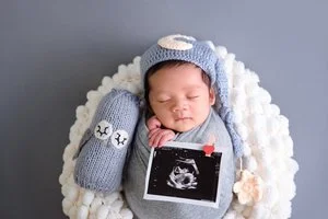 Sleeping newborn in a grey swaddle and moon-decorated knitted hat in a white-lined basket beside a knitted rabbit toy and an ultrasound picture