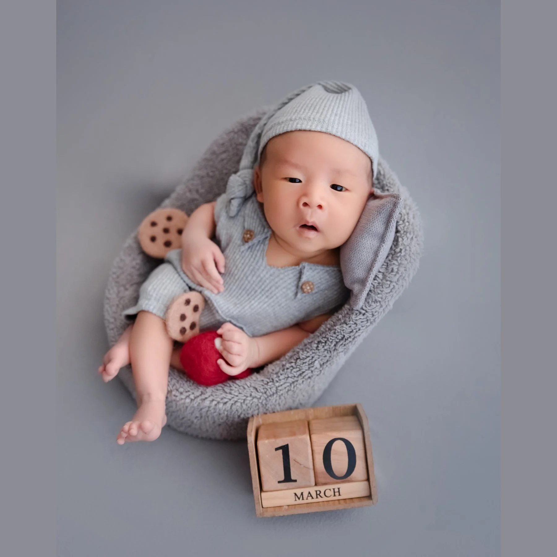 Awake newborn in a grey knitted hat and romper reclining in a soft grey basket with a red toy cup, miniature cookies and wooden number blocks