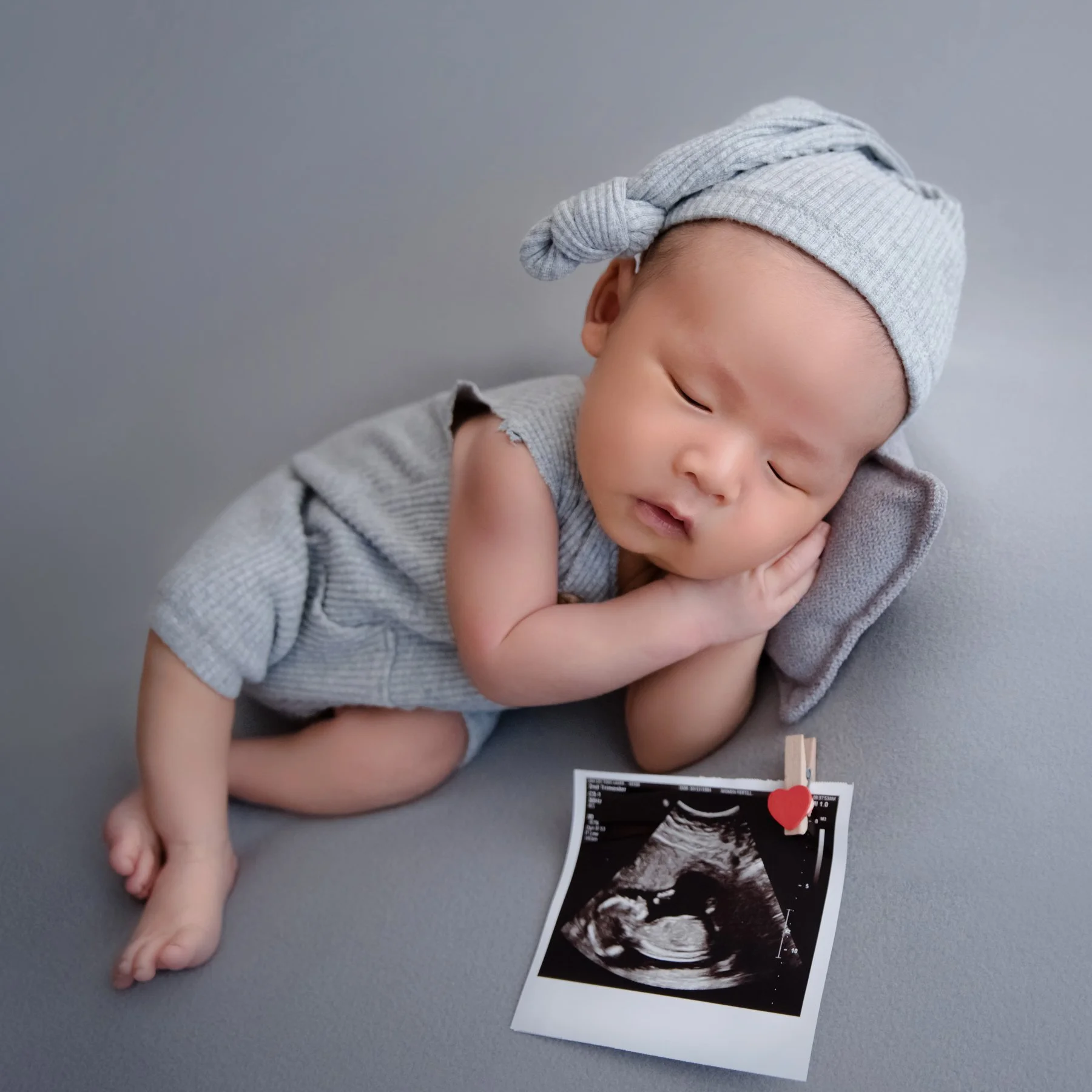 Sleeping newborn in a grey knitted bonnet and outfit curled on one side with hands beside the cheek near an ultrasound picture on a grey backdrop