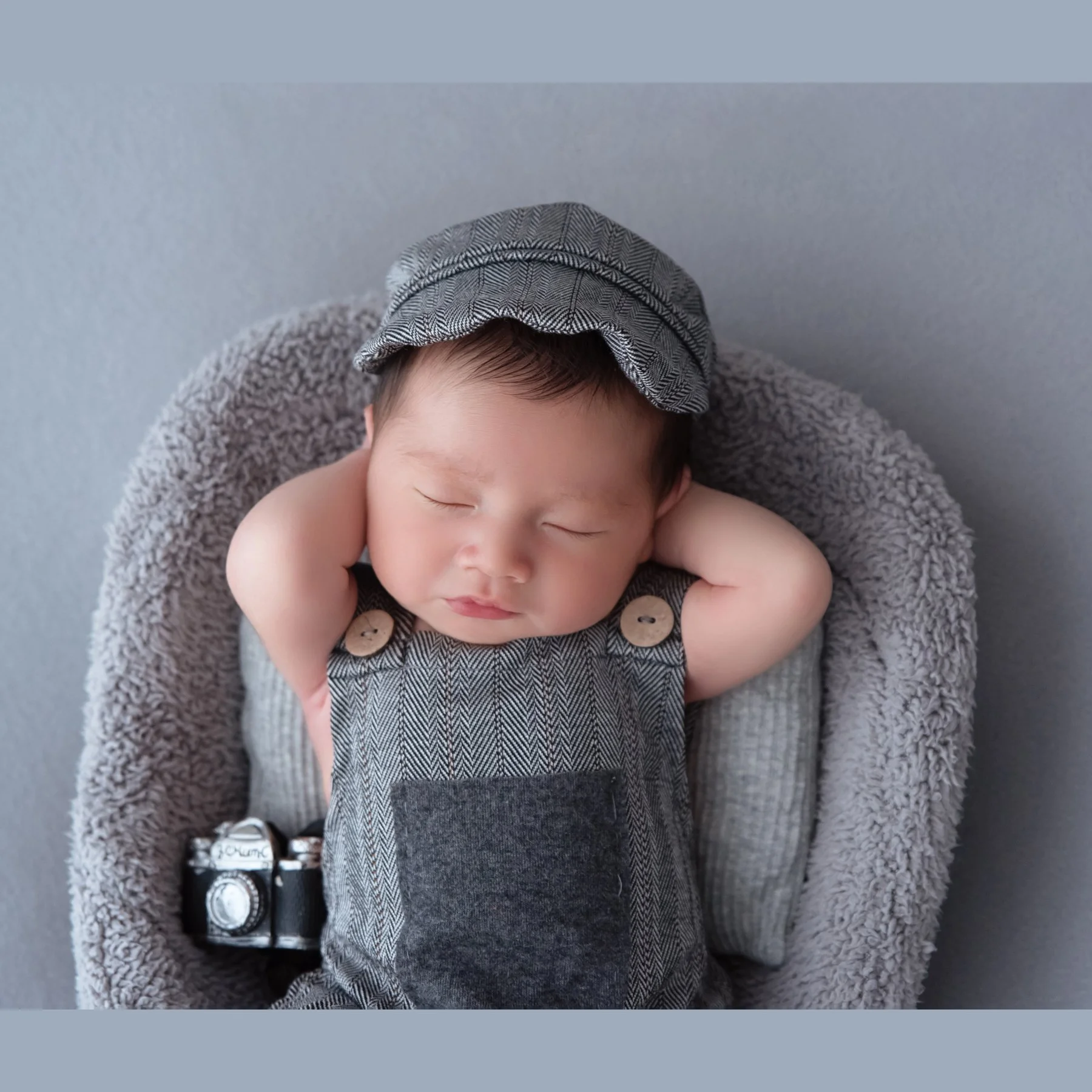 Close-up of a sleeping newborn in a grey cap and knitted overalls with both hands behind the head and a miniature camera beside the body