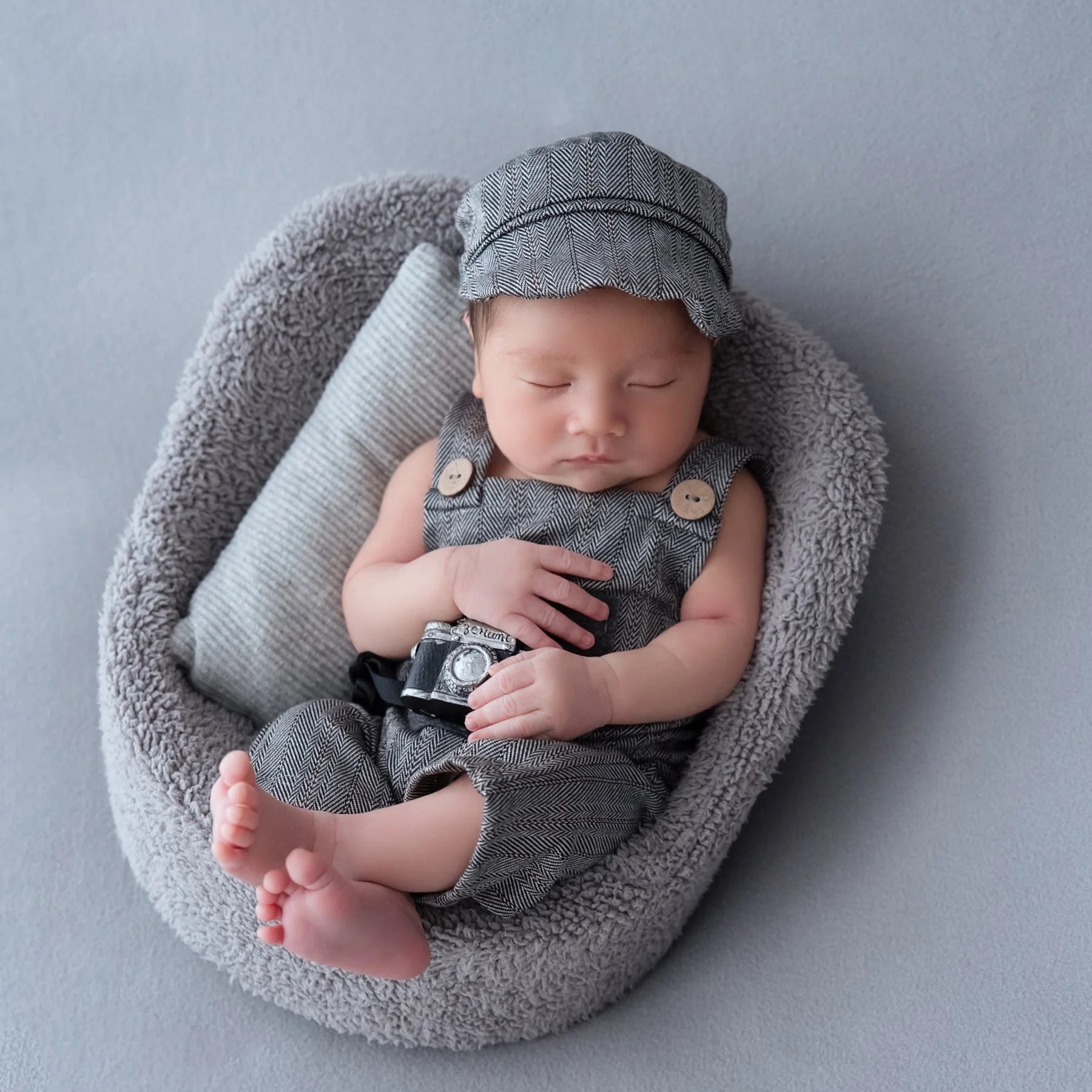 Sleeping newborn in a grey cap and knitted overalls holding a miniature camera while reclining in a grey basket
