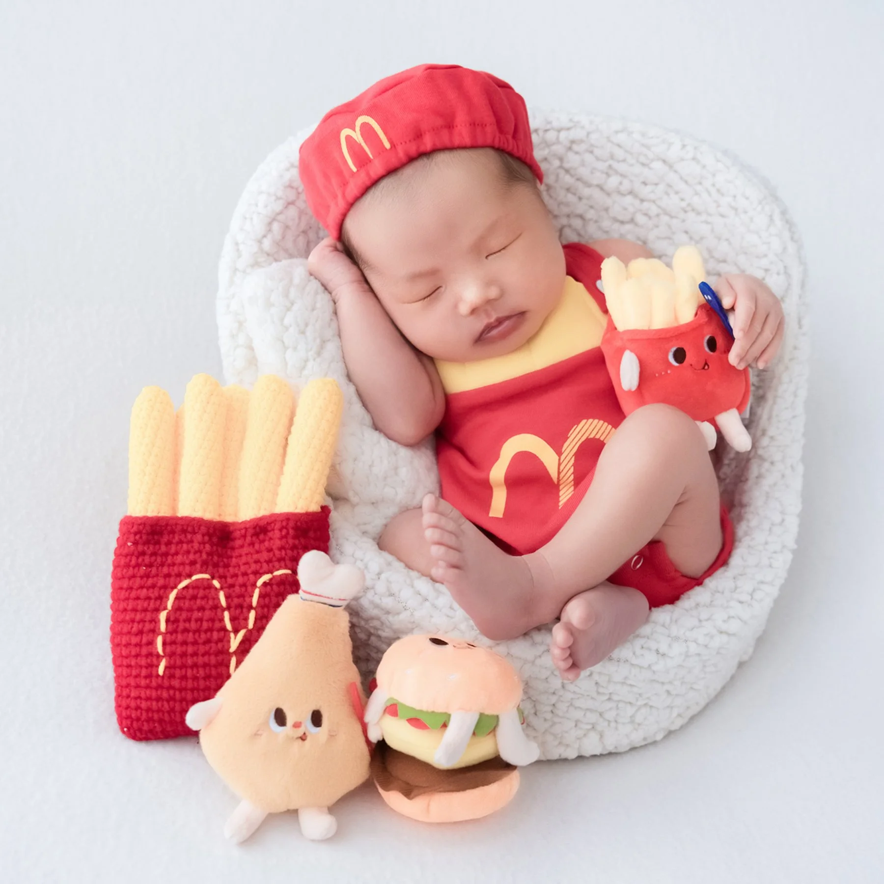 Sleeping newborn in a red McDonald’s cap and fries outfit in a white basket, surrounded by fast-food plush toys