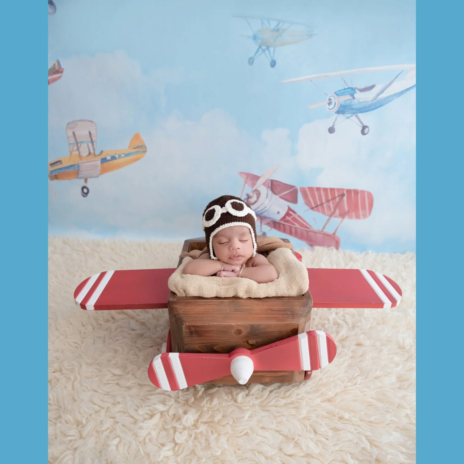 Sleeping newborn in a brown aviator hat and goggles resting on folded arms in a wooden airplane prop with red wings