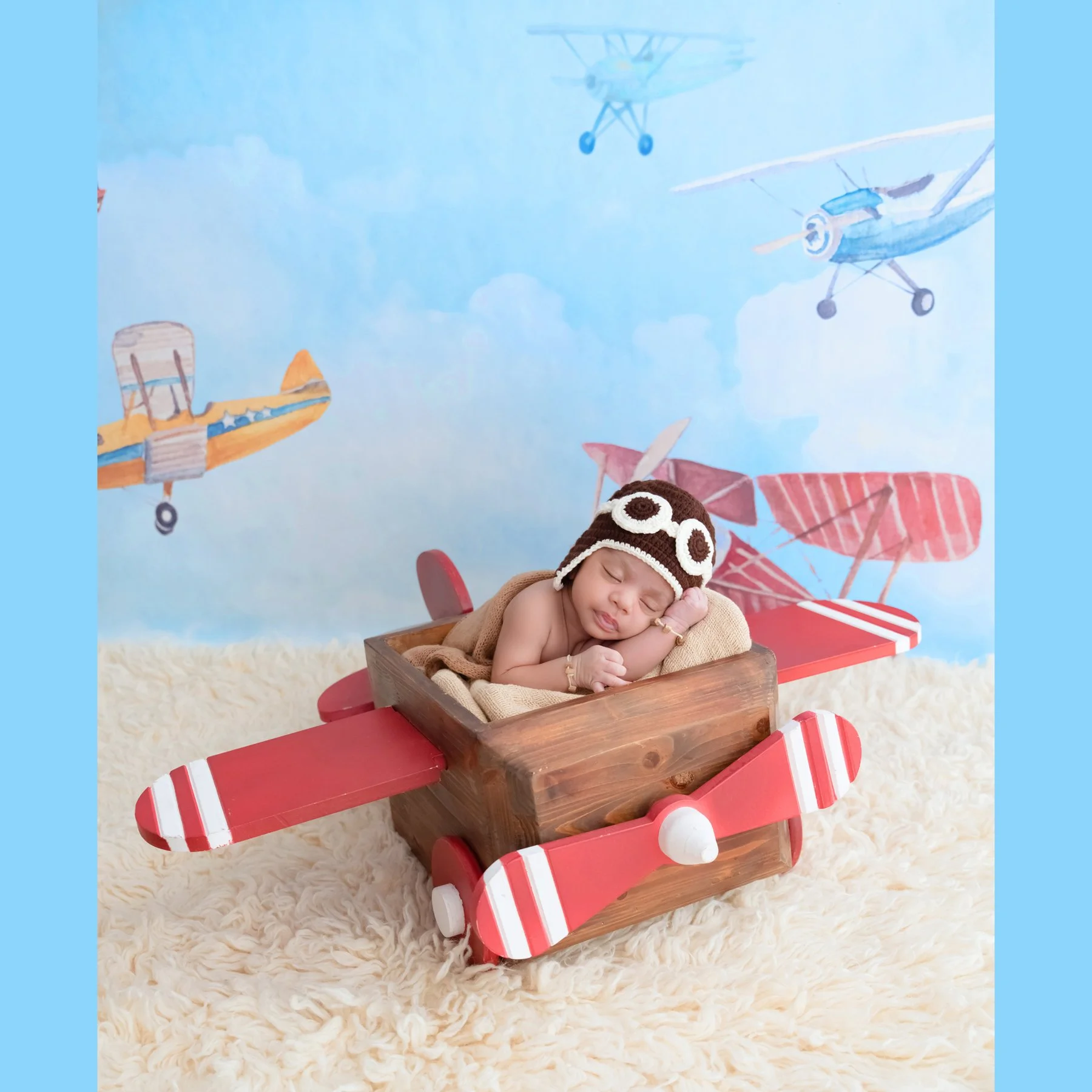 Sleeping newborn in an aviator hat and goggles resting one cheek on folded arms in a red-winged wooden airplane against a vintage aircraft backdrop