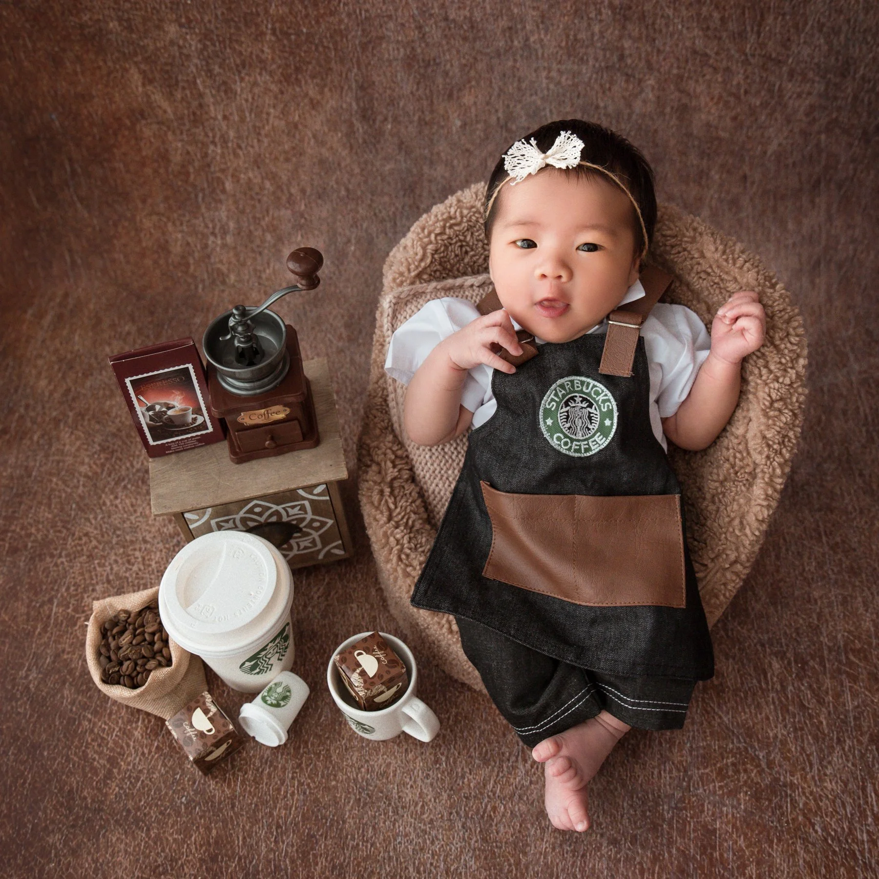 Awake newborn wearing a floral headband and Starbucks barista apron in a woven basket beside a coffee grinder and cups
