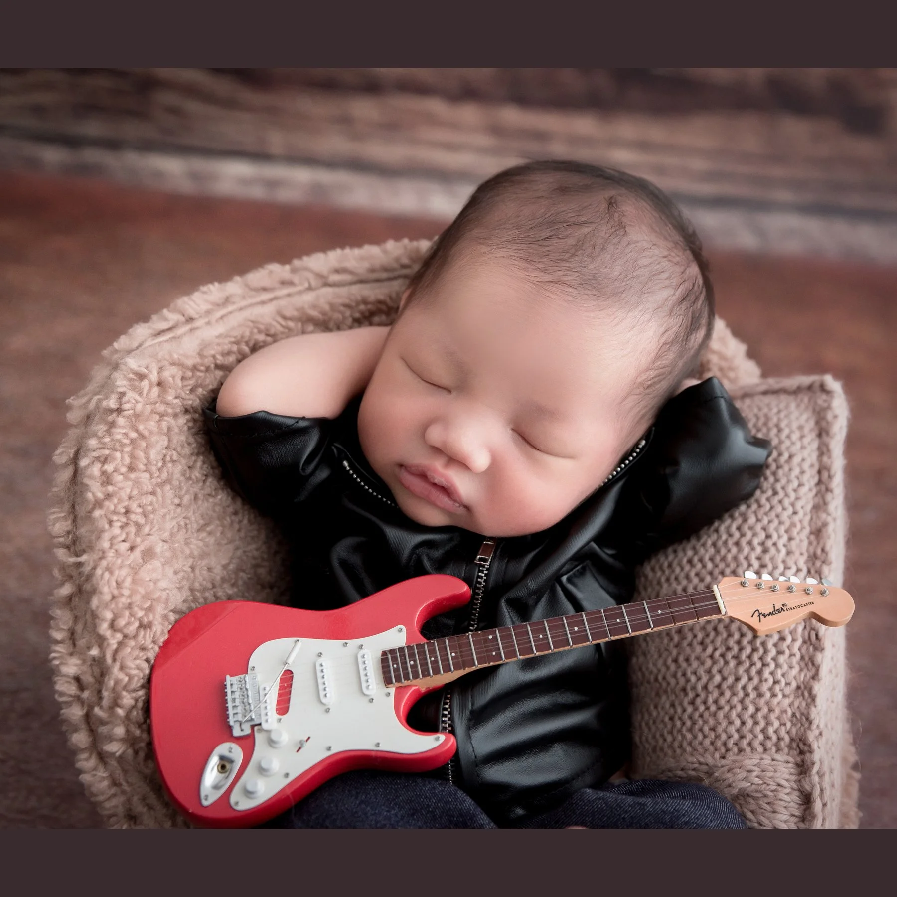 Close-up of a sleeping newborn in a black jacket curled in a beige basket with a red toy electric guitar