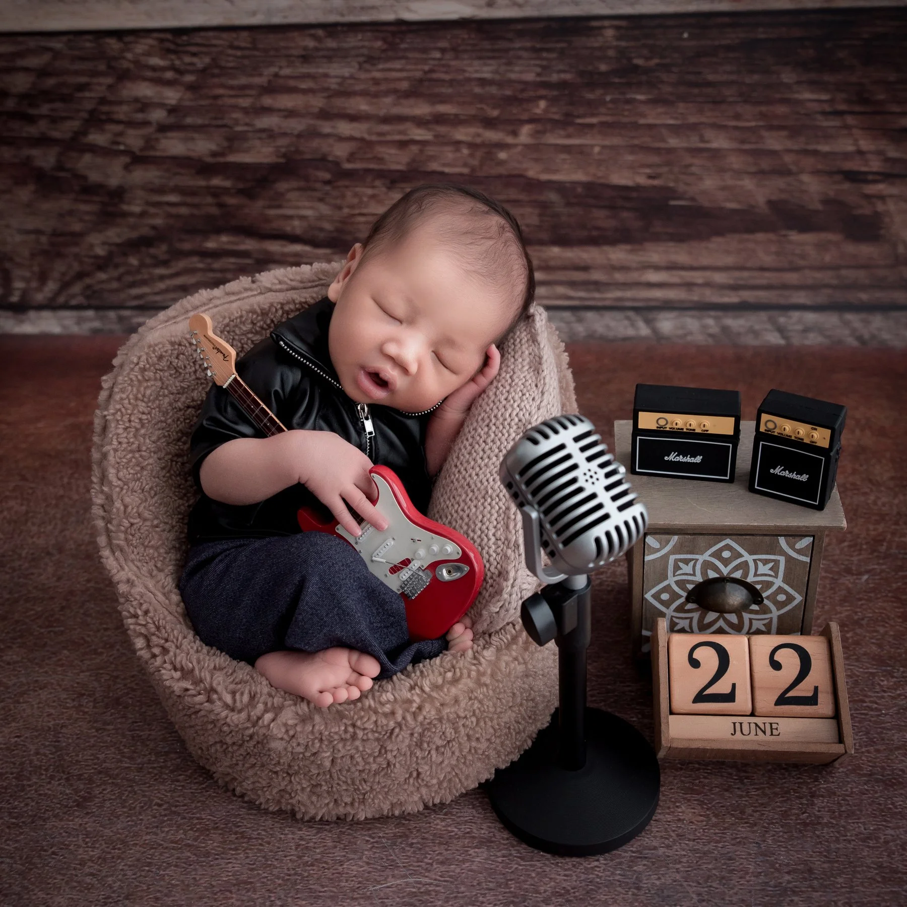 Sleeping newborn in a black jacket curled in a beige basket with a red toy guitar beside a microphone and small amplifier