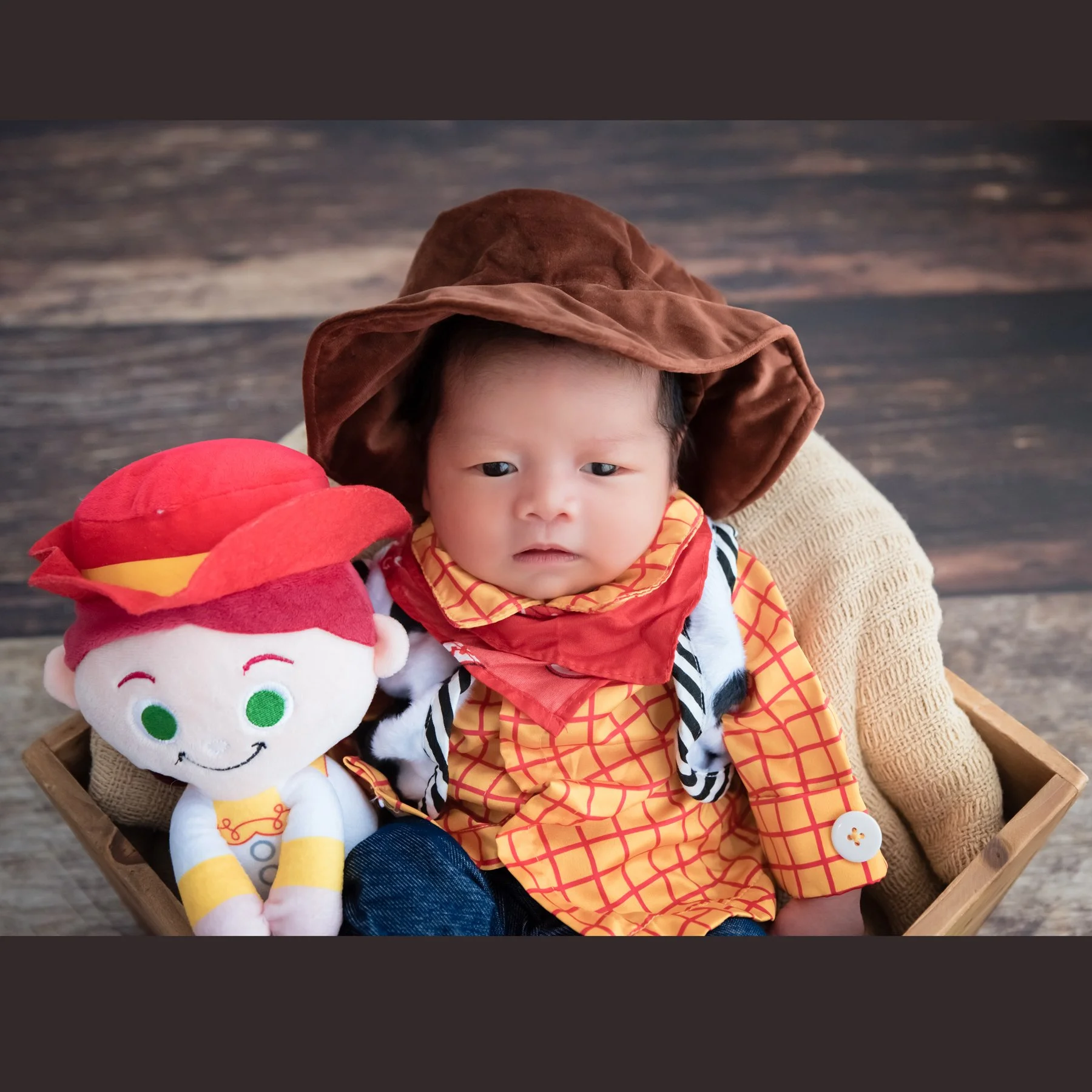 Awake newborn wearing a brown cowboy hat and Woody outfit with a Jessie plush beside the face