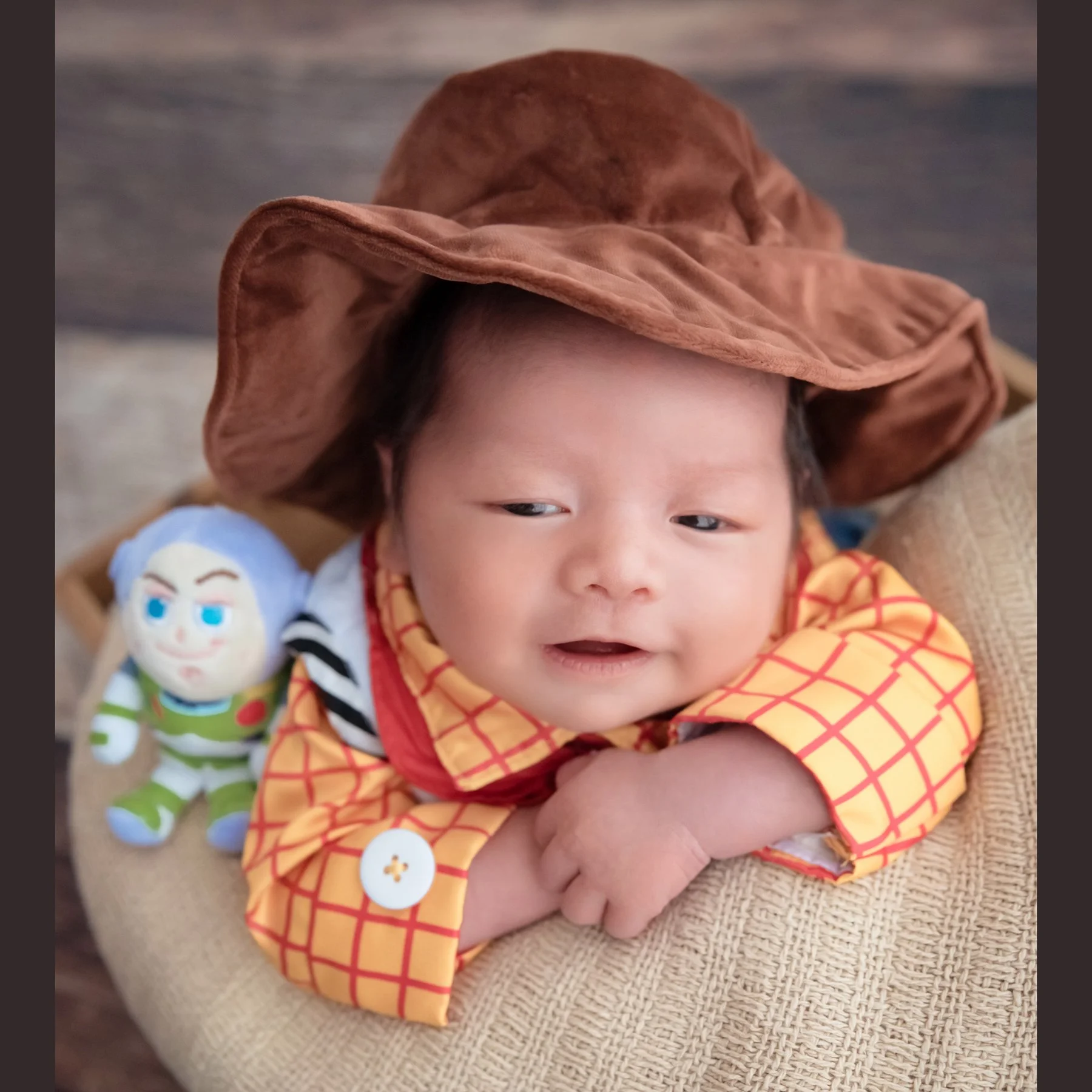 Awake newborn in a brown cowboy hat and Woody shirt resting on folded arms beside a Buzz Lightyear toy