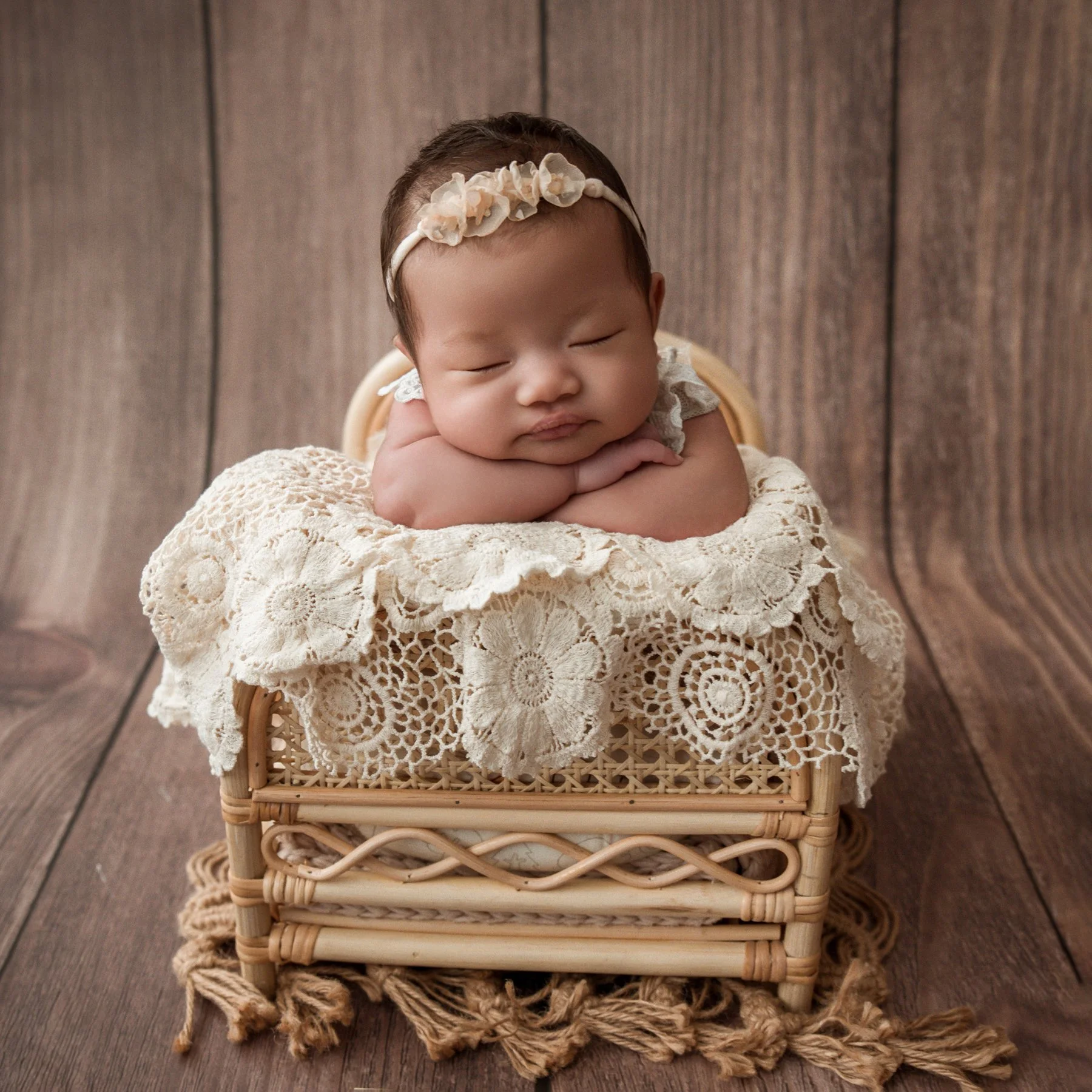 Sleeping newborn wearing a cream floral headband with chin resting on folded arms in a lace-covered wicker chair against a wooden backdrop