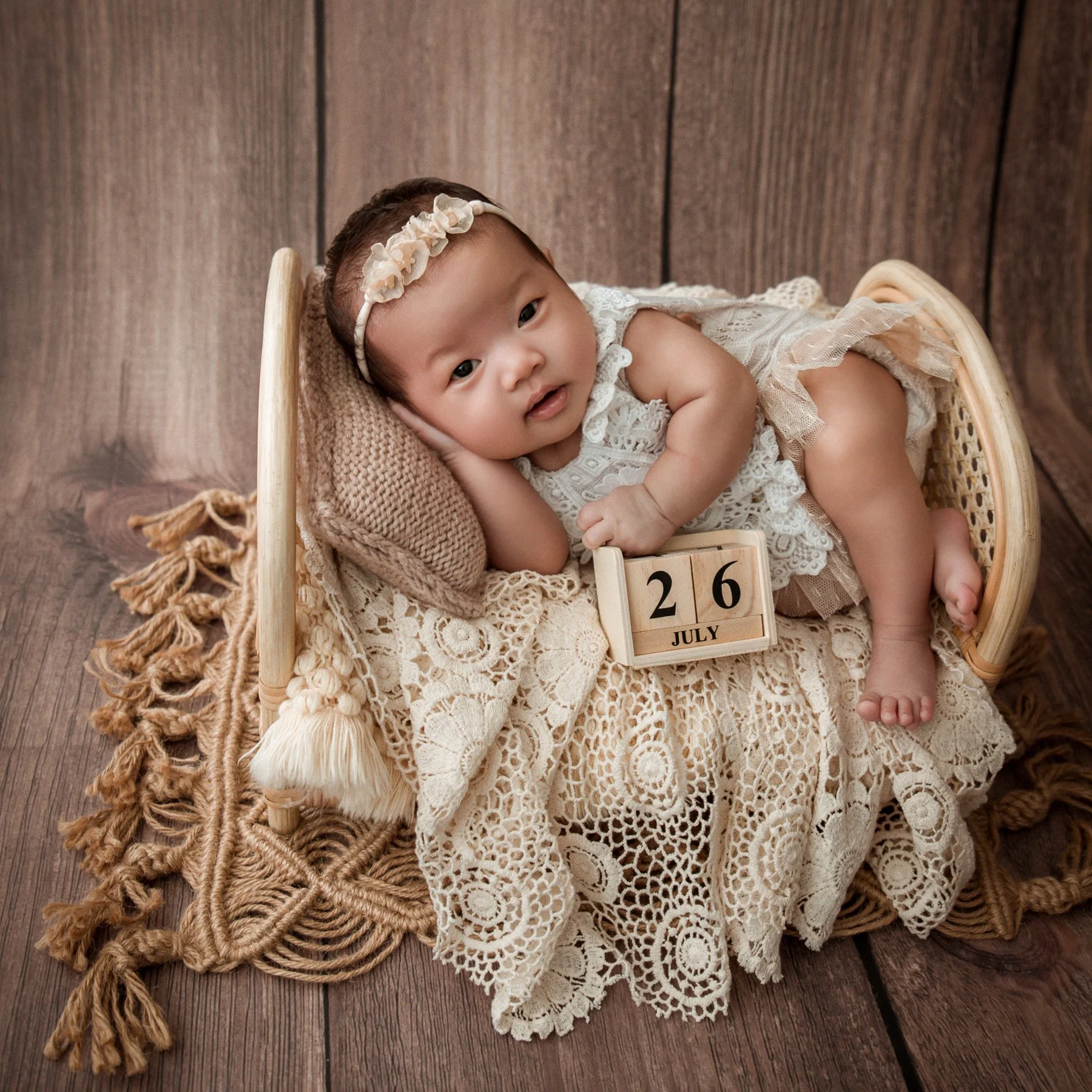 Awake newborn wearing a cream floral headband and lace dress reclining sideways on a lace-covered wicker chair against a wooden backdrop