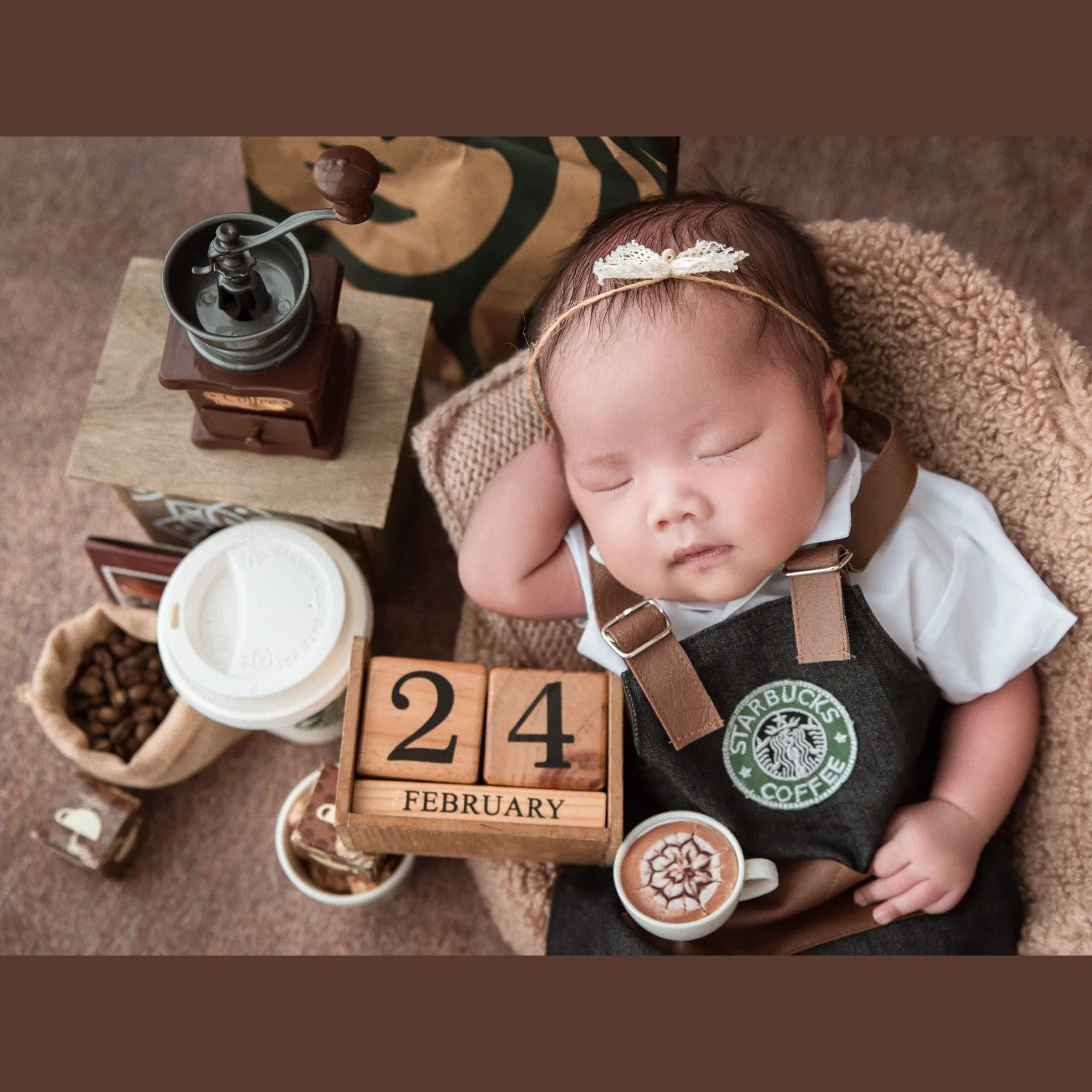 Sleeping newborn wearing a floral headband and Starbucks barista apron with one hand beside the cheek in a woven basket among coffee cups and a grinder