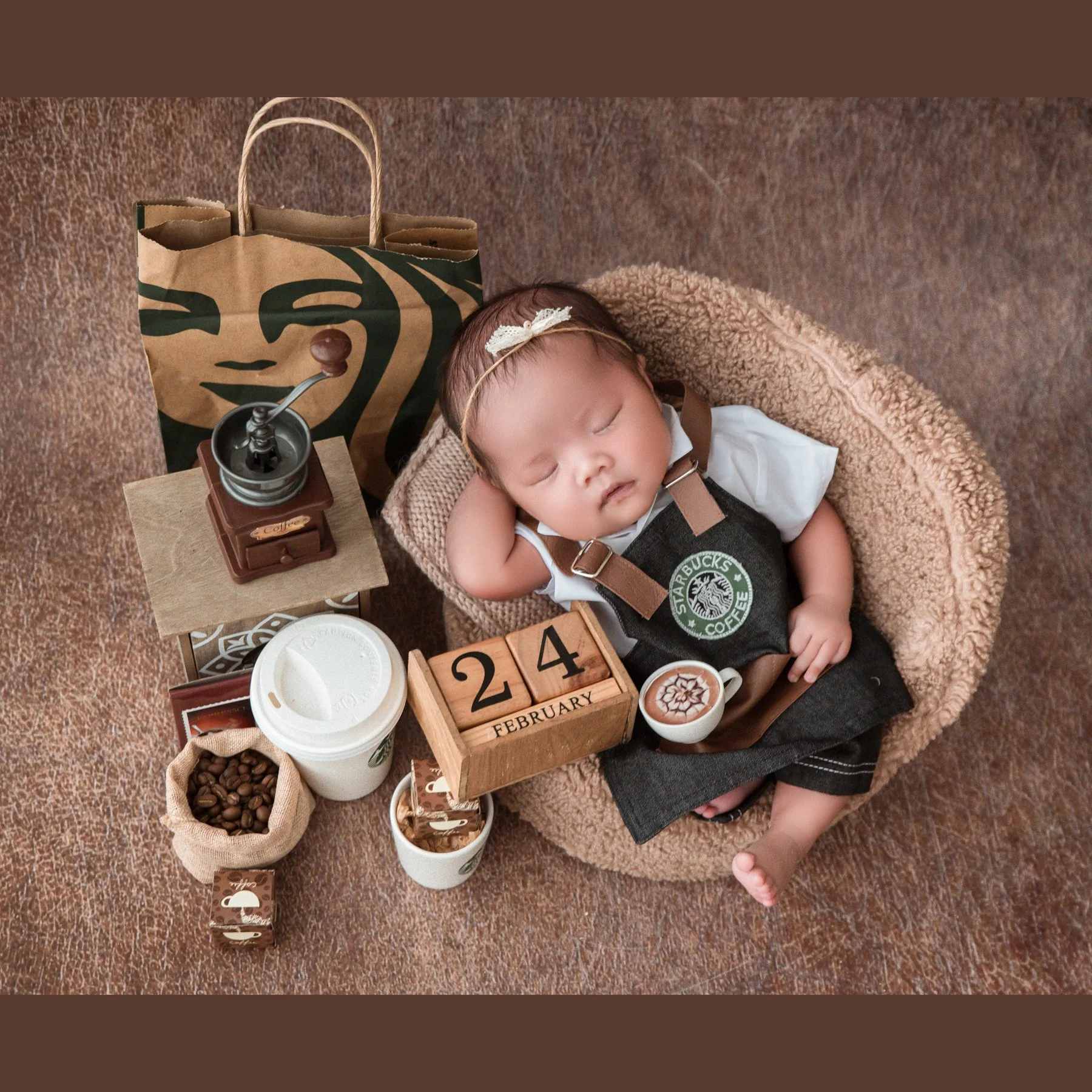 Sleeping newborn wearing a floral headband and Starbucks barista apron in a woven basket beside a coffee grinder, cups and wooden number blocks