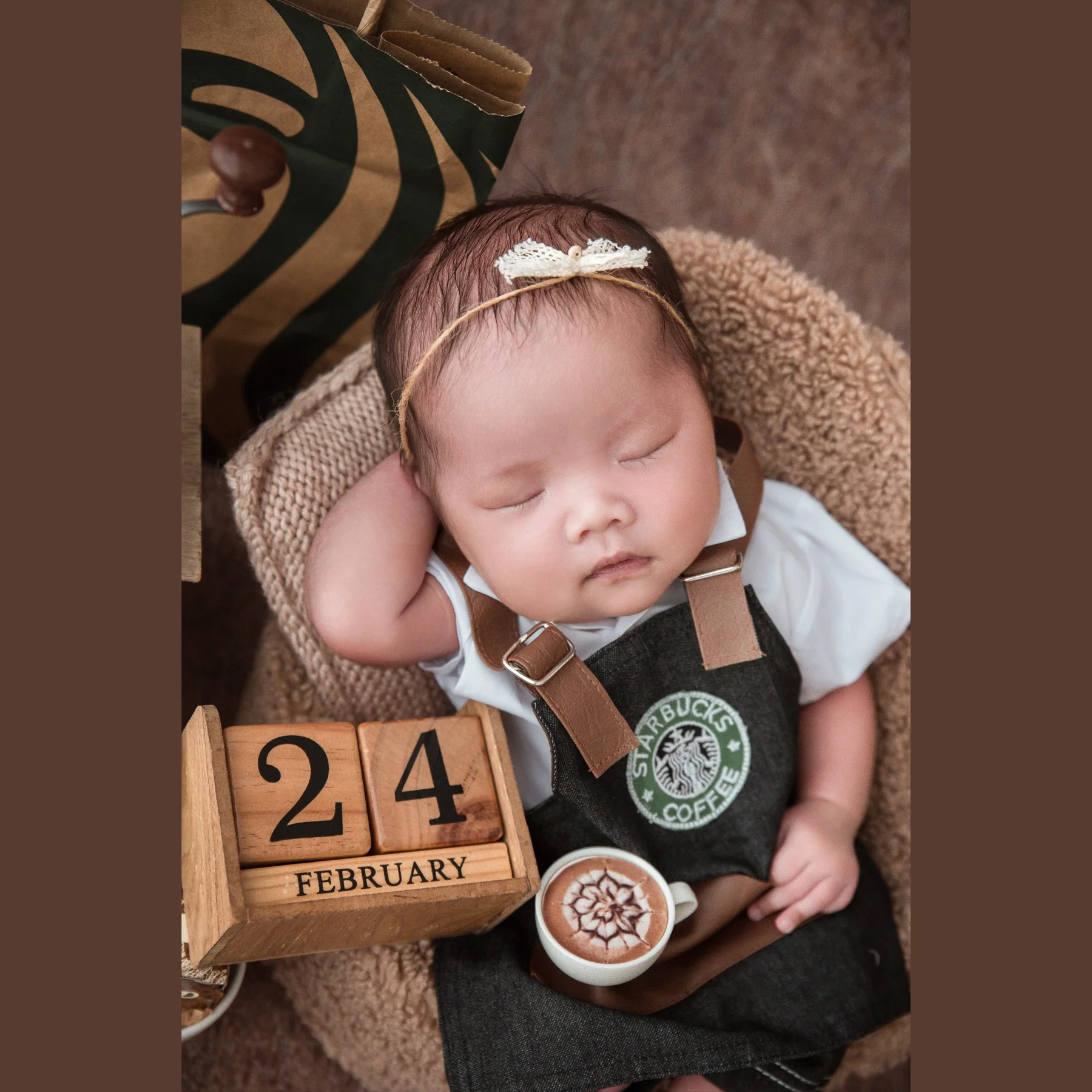 Close-up of a sleeping newborn wearing a floral headband and Starbucks barista apron beside wooden number blocks and coffee props