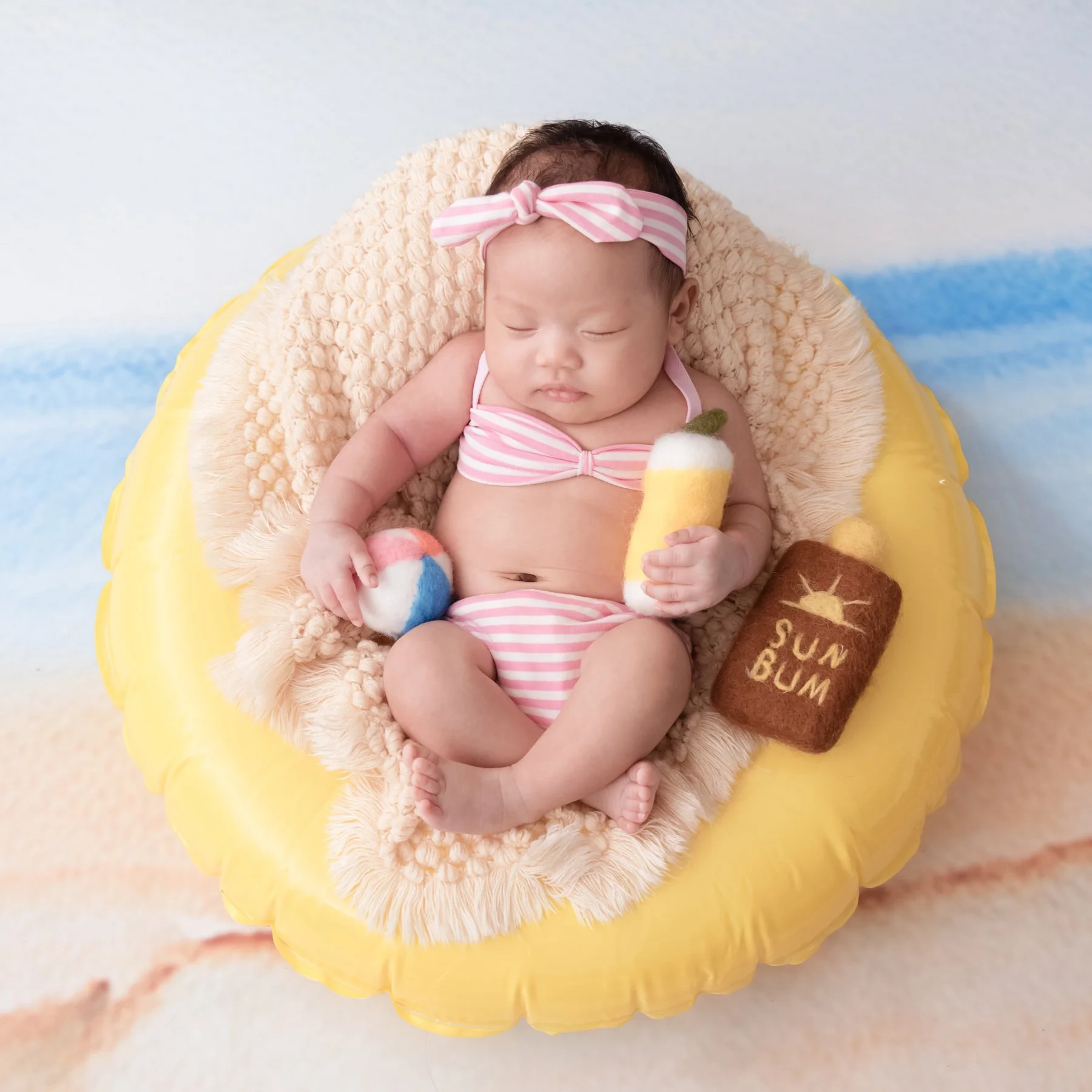 Sleeping newborn in a pink swimsuit and matching headband reclining in a yellow inflatable ring with miniature drink and beach-ball props