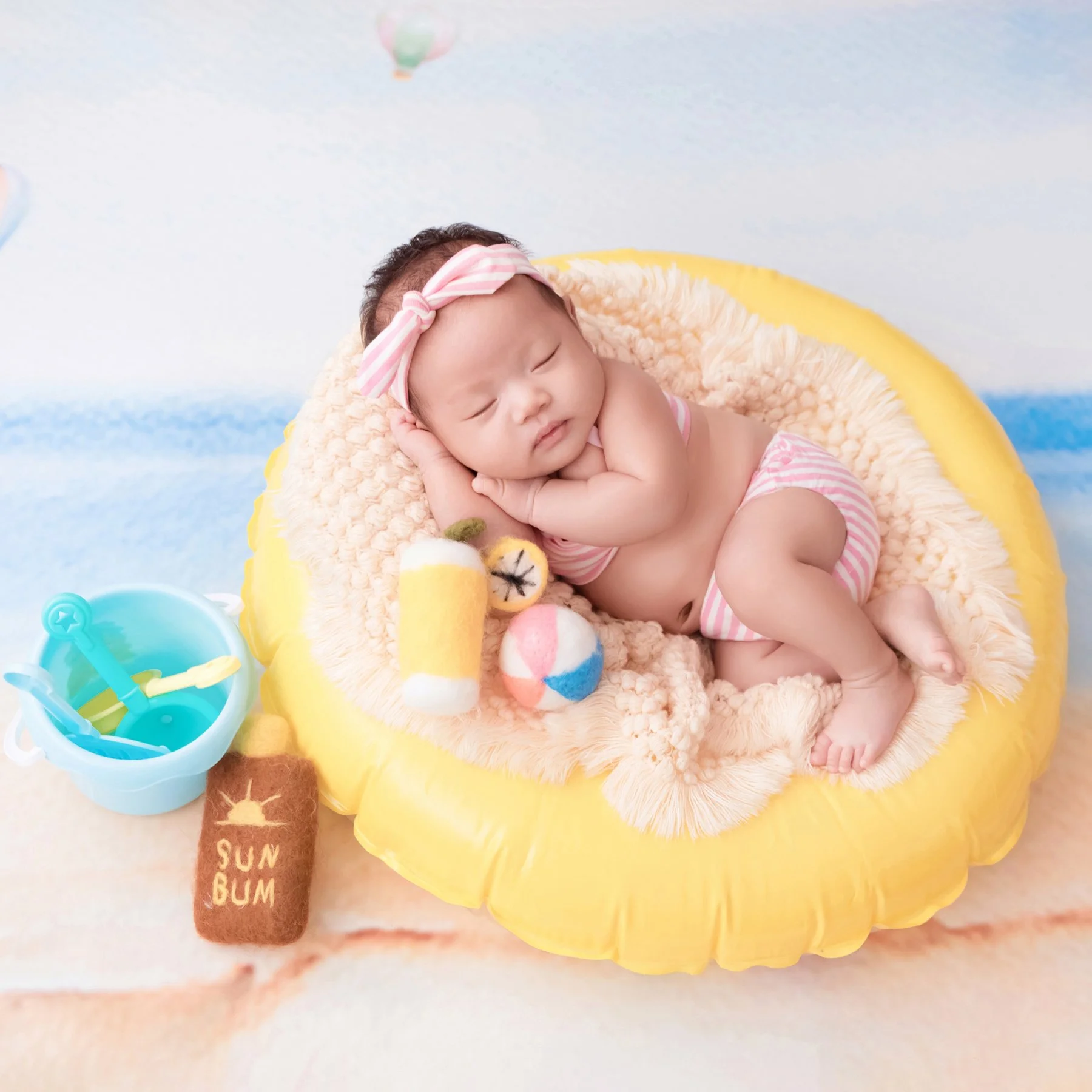 Sleeping newborn in a pink swimsuit and matching headband curled on one side in a yellow inflatable ring beside a toy drink and blue beach bucket