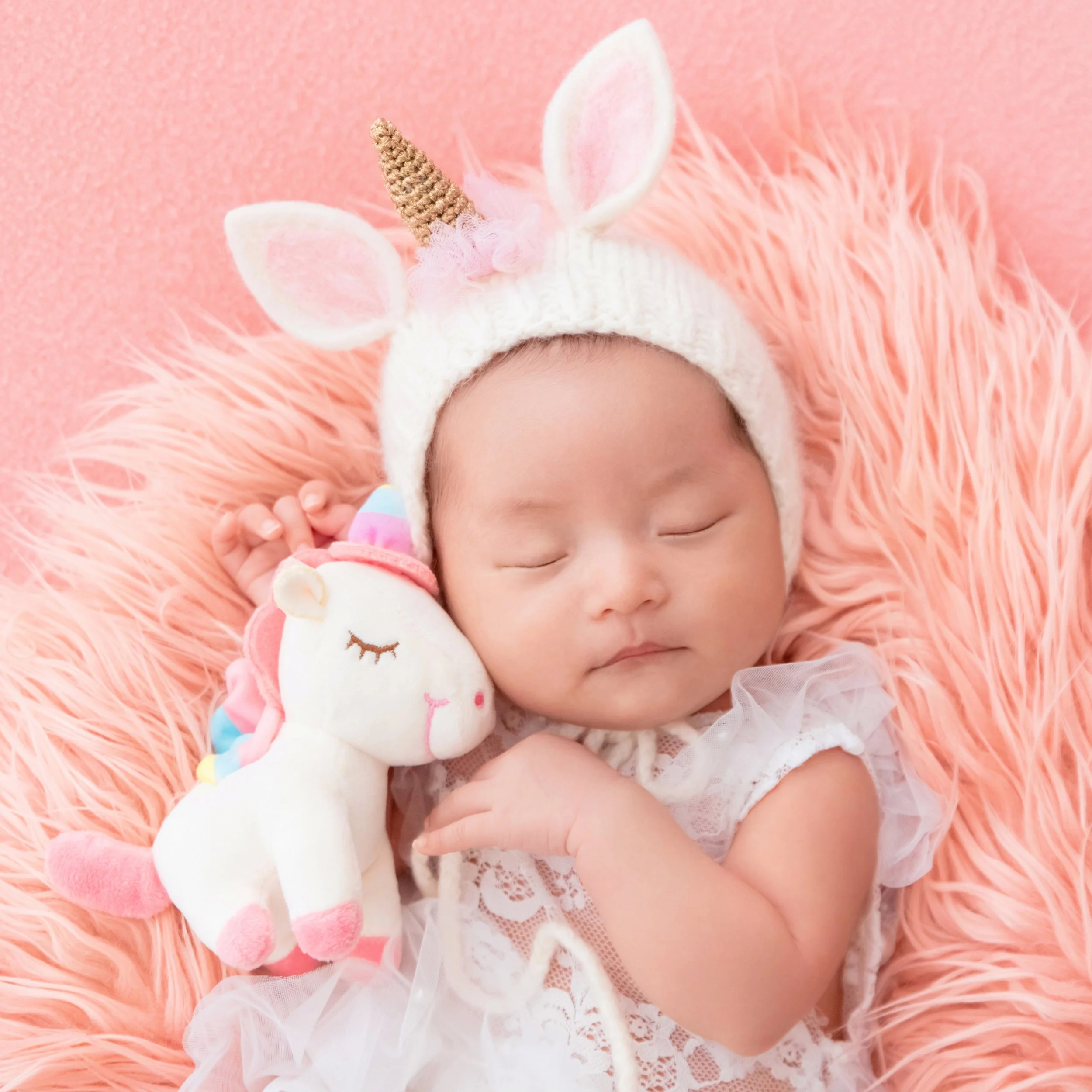 Sleeping newborn in a white unicorn hat and lace outfit nestled on pink fur beside a unicorn plush