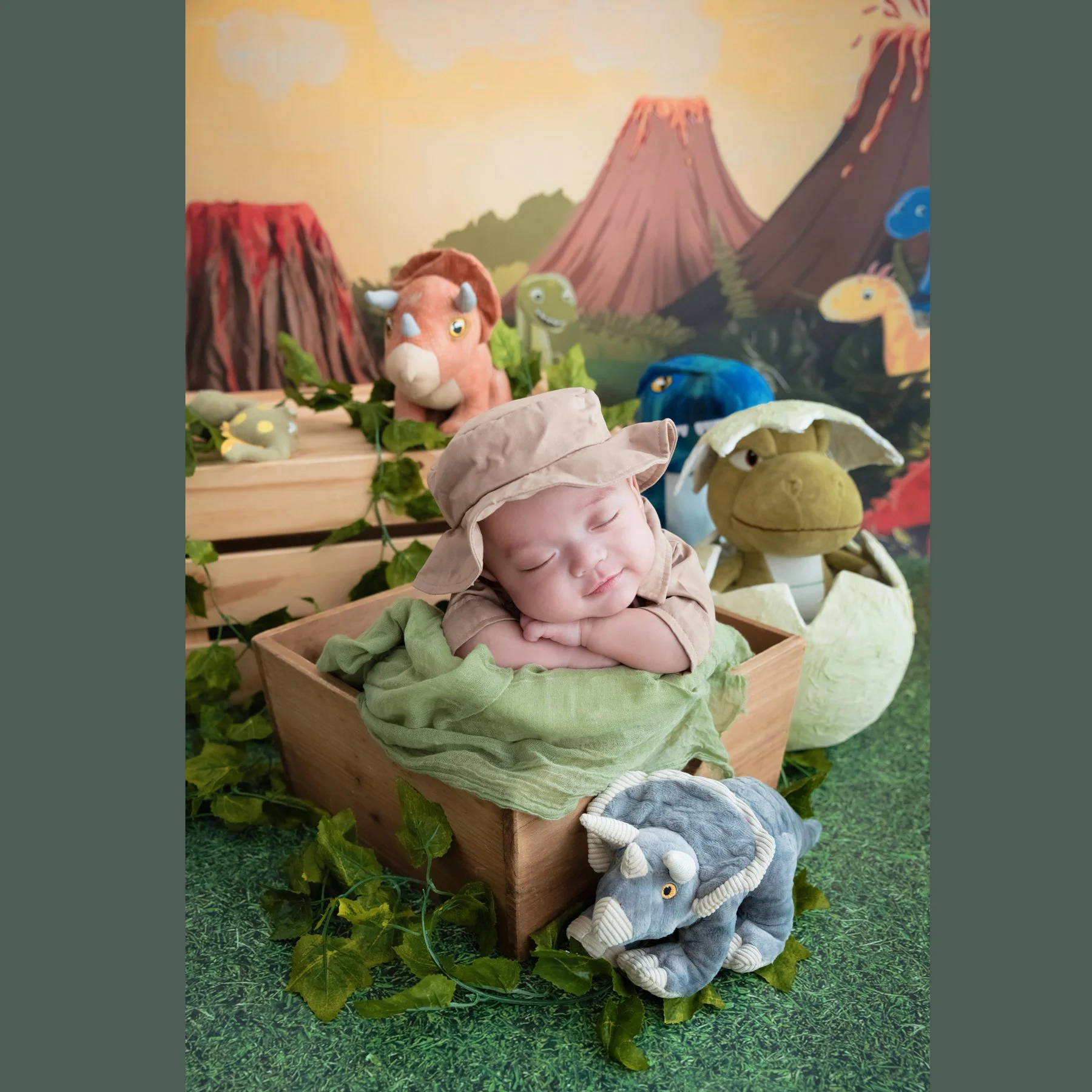 Sleeping newborn in a safari hat and green wrap resting on folded arms in a wooden crate among dinosaur toys against a volcano backdrop
