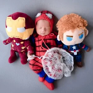 Sleeping newborn in a Spider-Man costume between Iron Man and Captain America plush toys with a white web prop