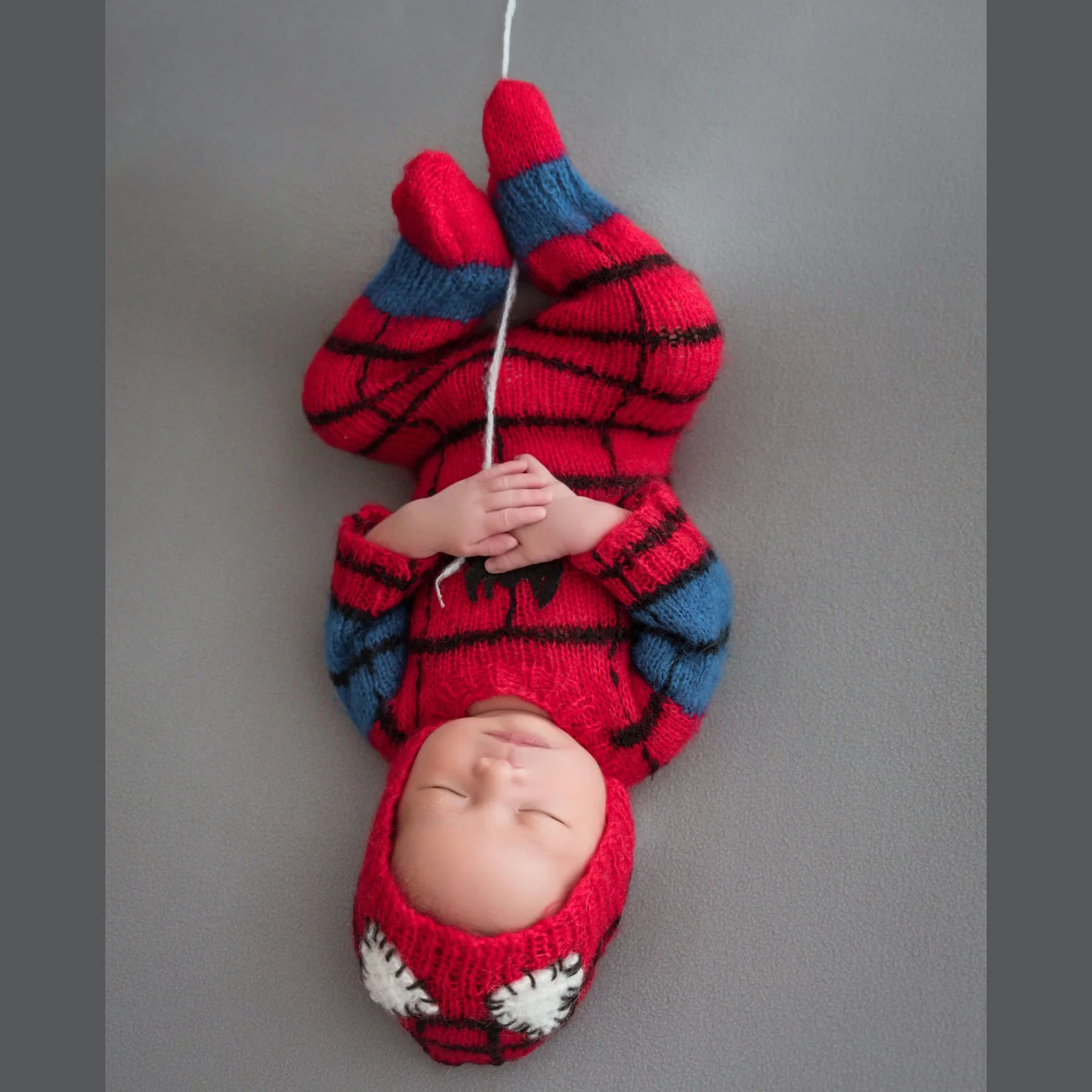 Sleeping newborn in a knitted Spider-Man costume shown upside down, holding a white string against a grey backdrop