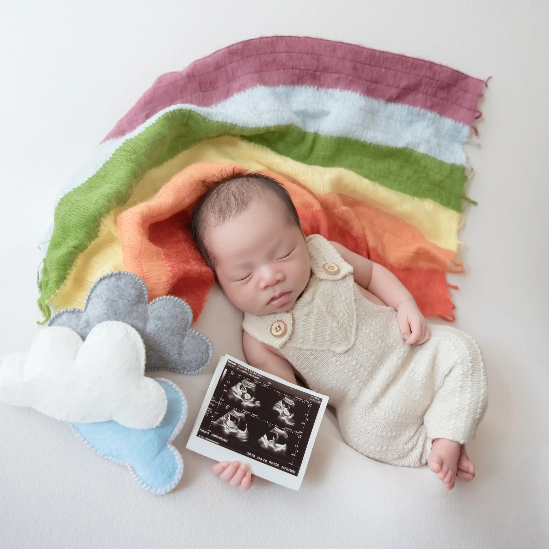 Sleeping newborn in cream knitted overalls reclining beneath colourful rainbow fabric beside felt clouds and an ultrasound picture
